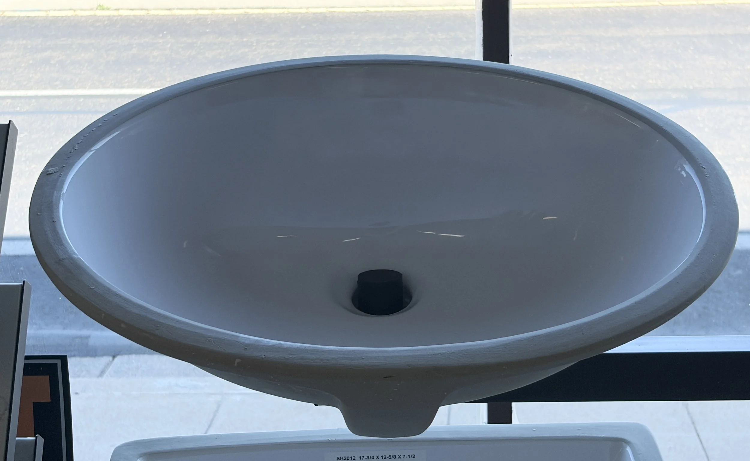 A white oval-shaped sink with a black drain stopper, mounted on a white countertop, with a window in the background showing an outdoor scene.