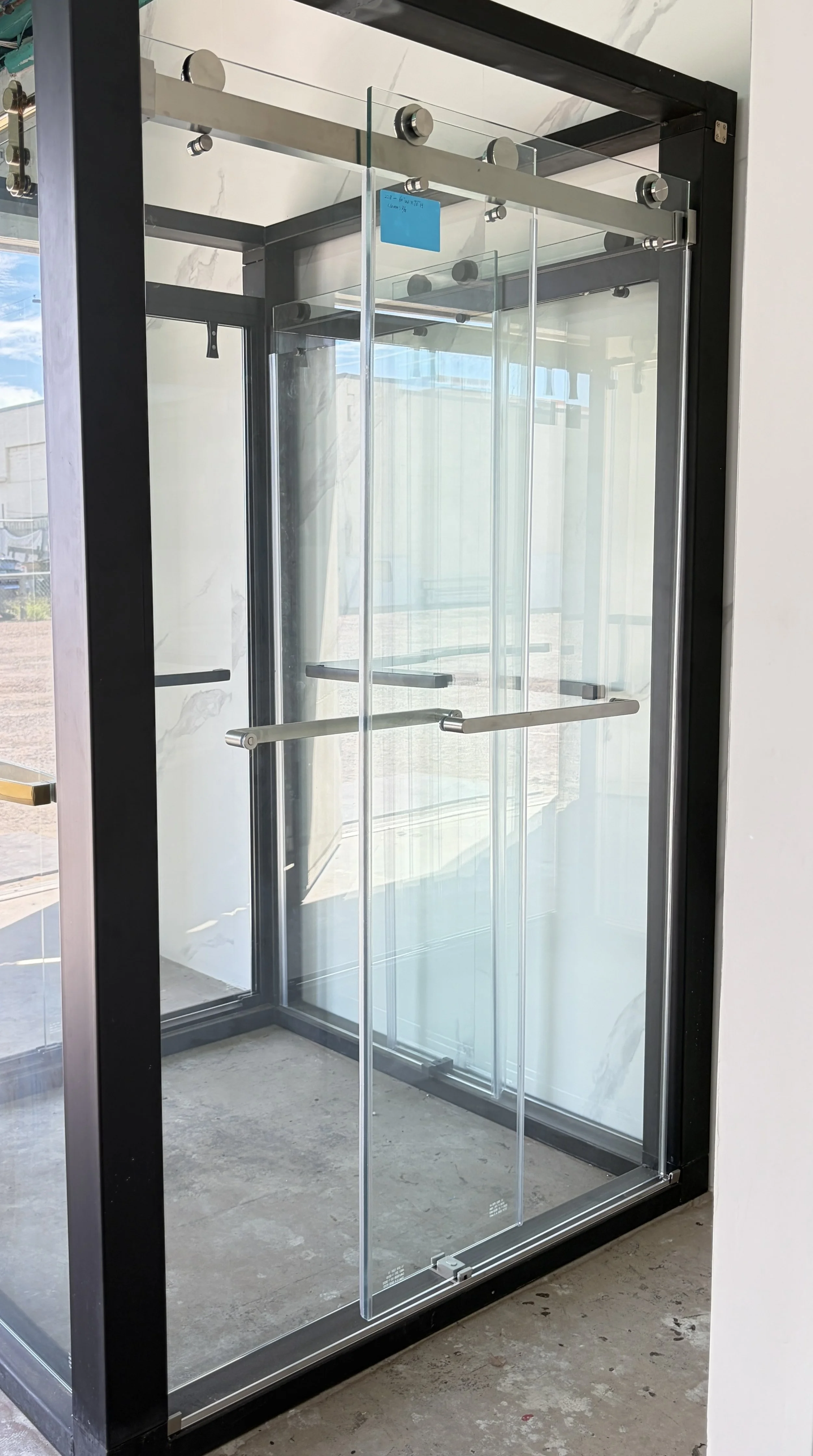 A modern glass and metal door with sliding mechanism and black frame, installed in a building with concrete floor and large windows.