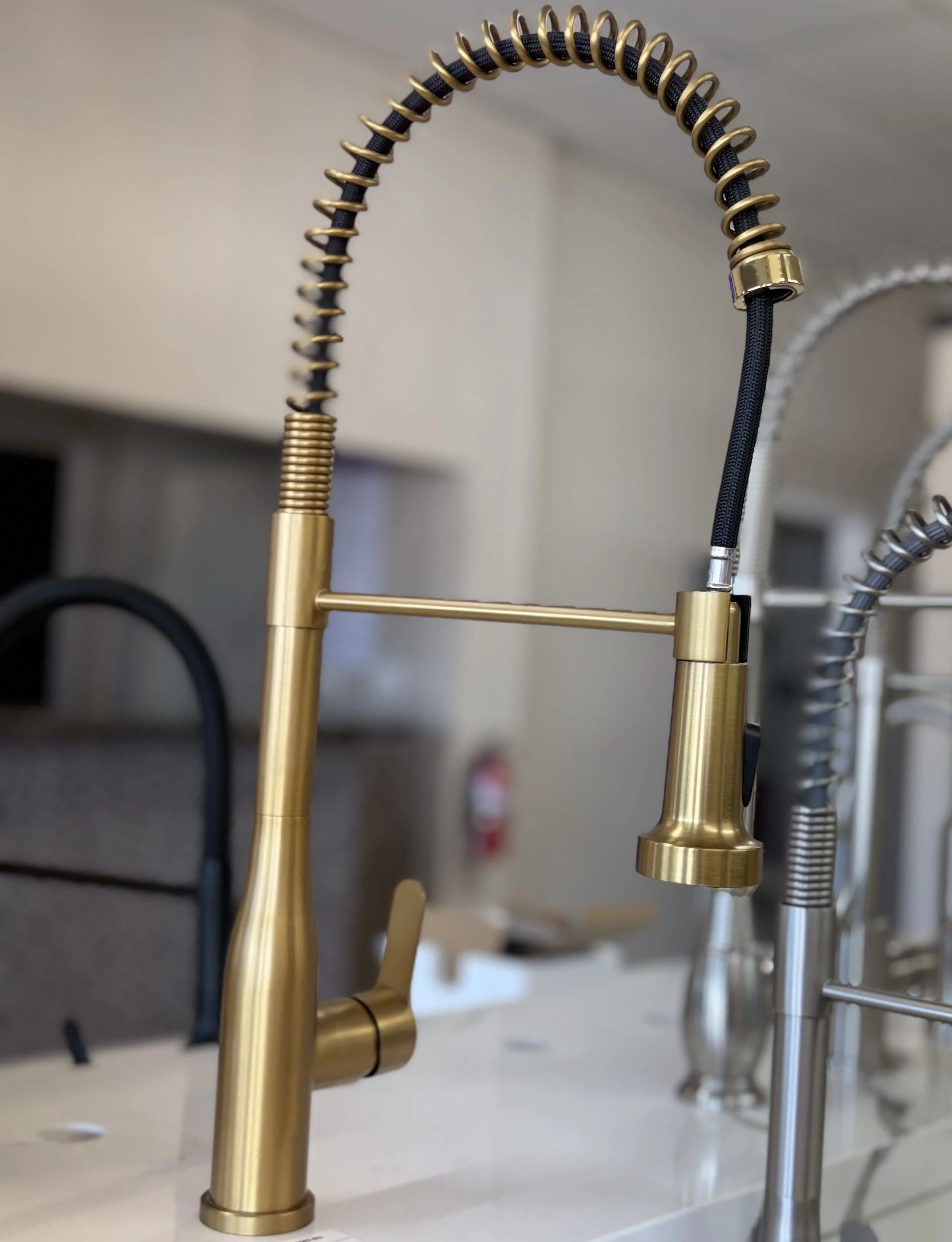 A close-up of a modern kitchen faucet with a flexible, coiled spring neck in a gold tone, with a black handle and a black hose. Other silver-colored faucets are visible in the background.