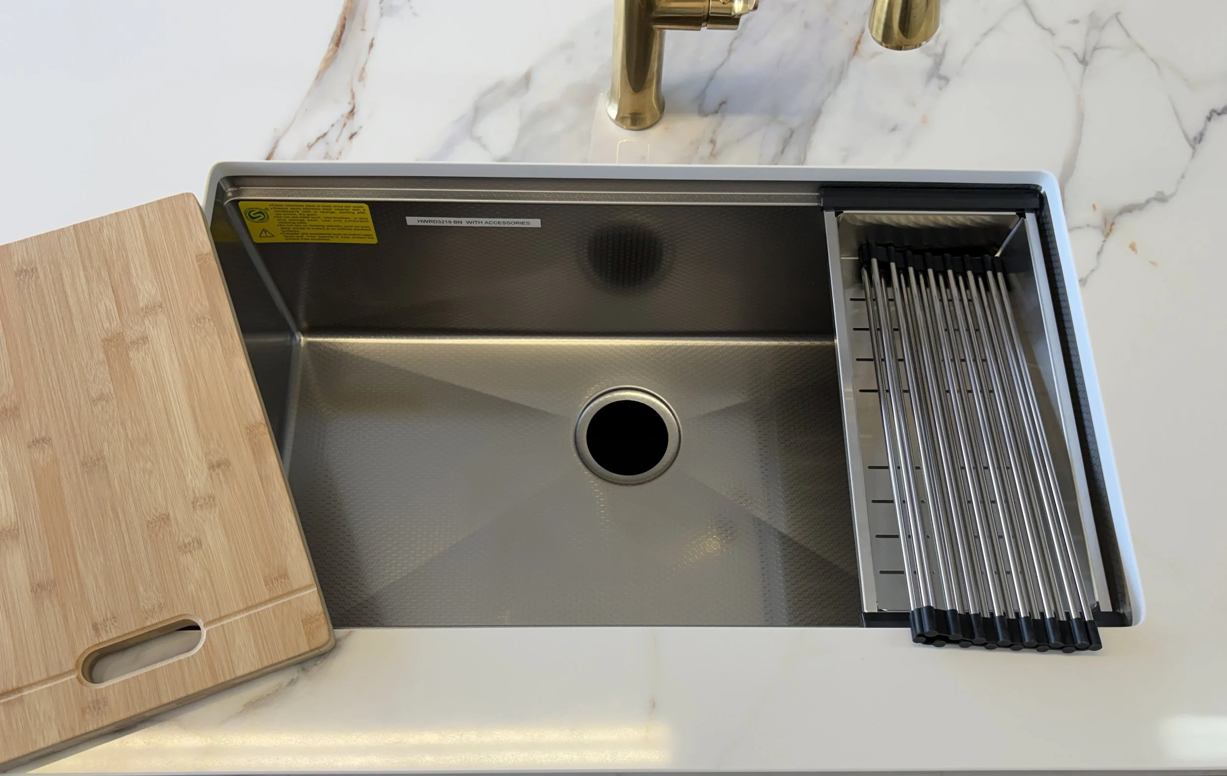 A stainless steel kitchen sink with a drain, silver rails on the right, a brass faucet above, and a bamboo cutting board on the left on a white marble countertop.