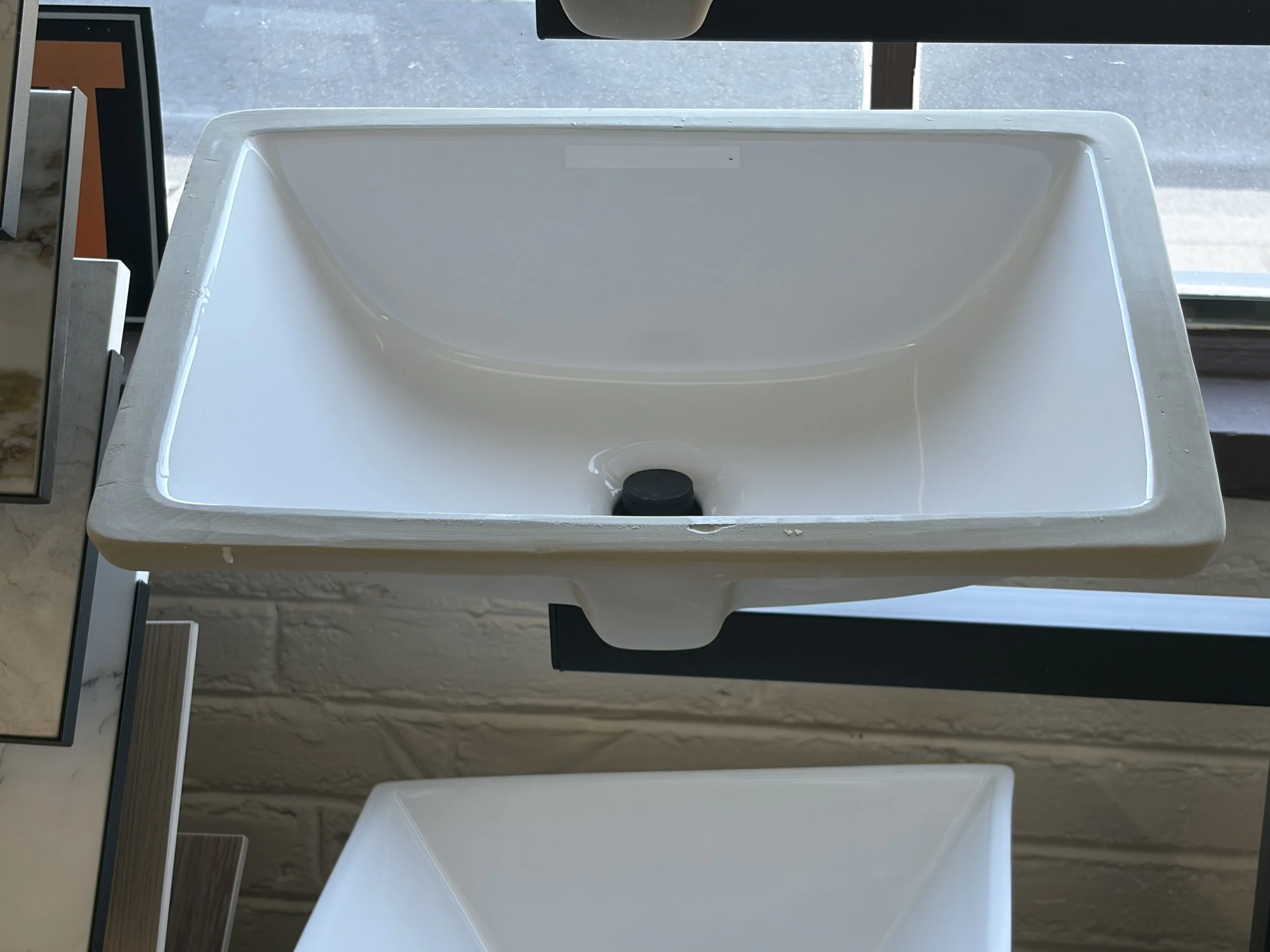 A white ceramic sink with a black drain stopper, placed on a countertop near large windows with a view of the outside.