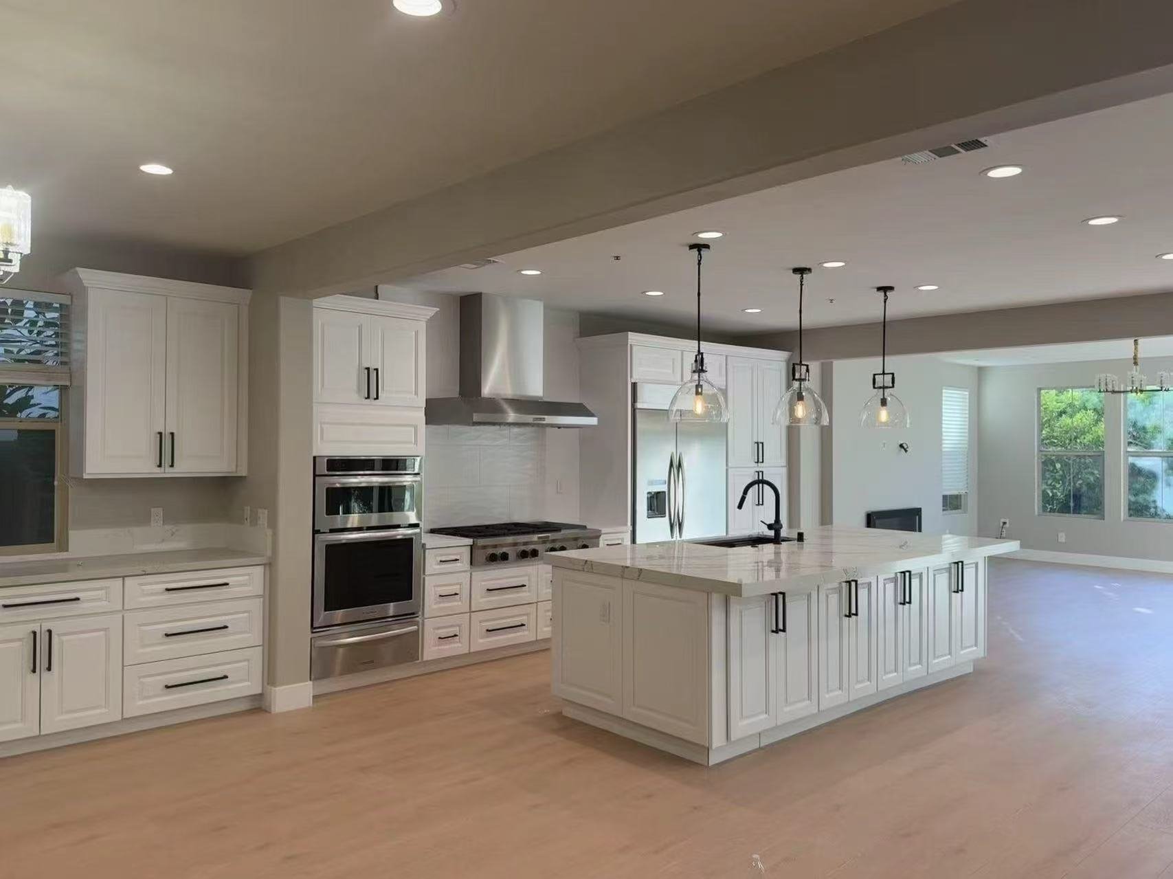 Modern kitchen with white cabinets, stainless steel appliances, and a large island with a marble countertop. Pendant lights hang above the island. There are large windows letting in natural light.