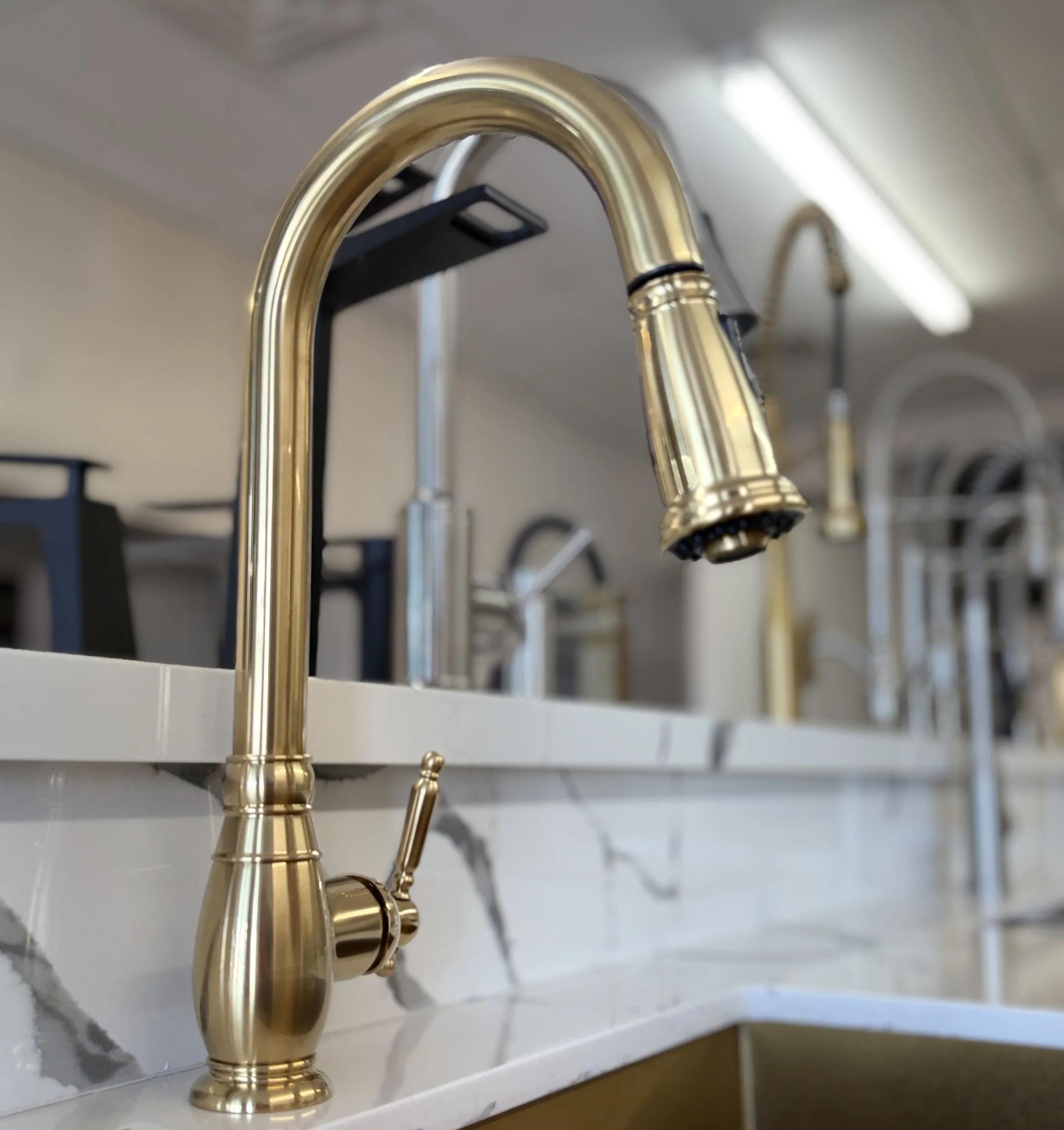 Golden kitchen faucet mounted on a white countertop with a sink. The background shows a blurred light-colored wall and other fixtures.