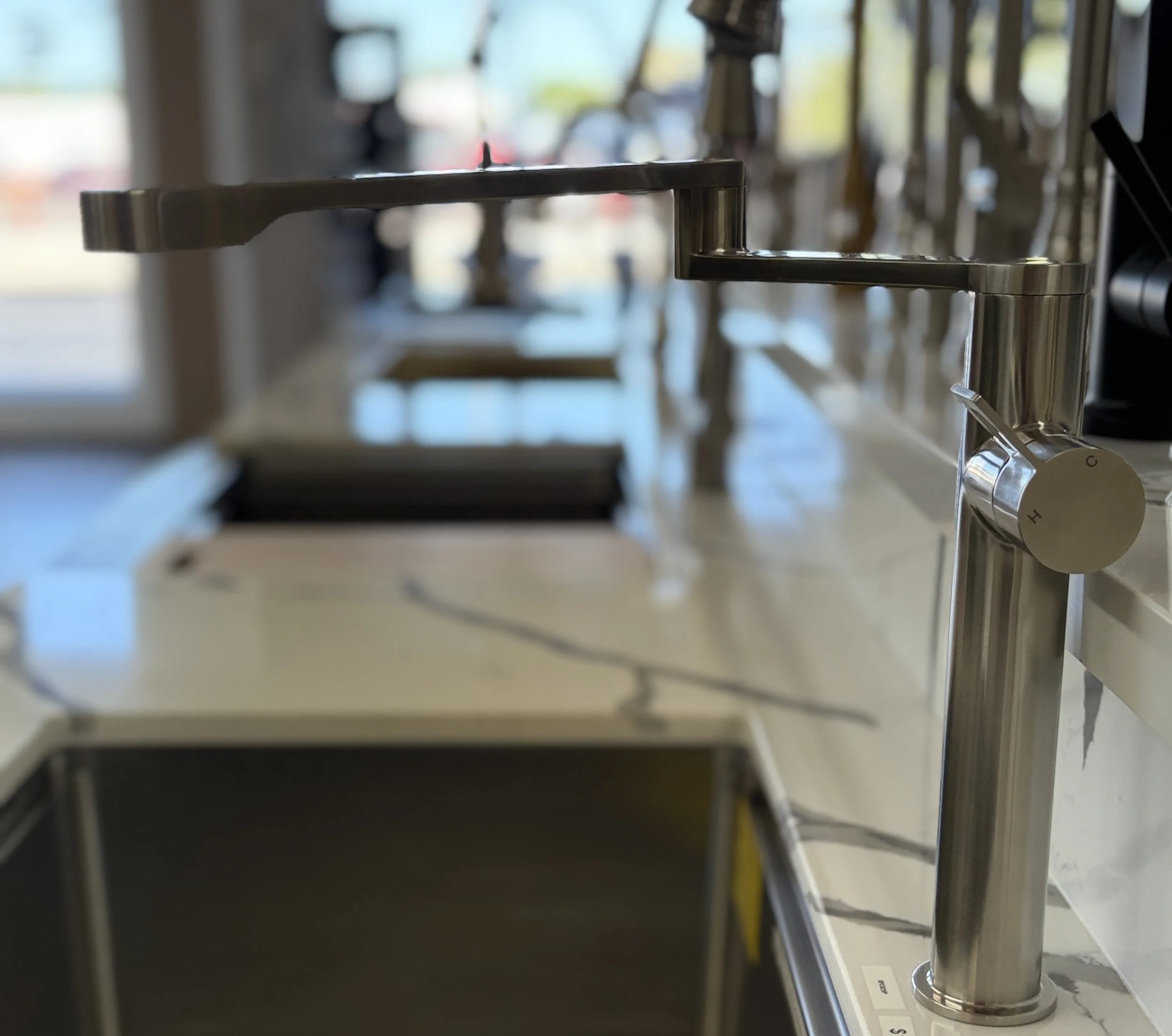 Close-up of a modern faucet with a background of multiple faucets and blurred outdoor scenery visible through a window.