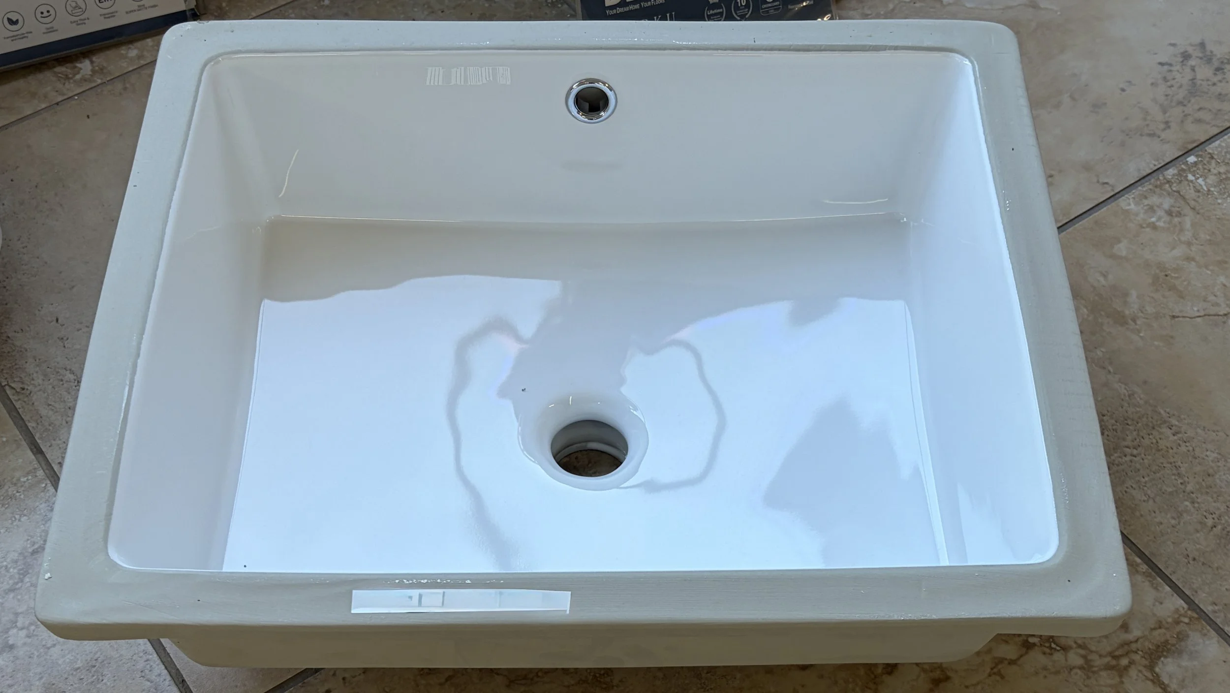 A white ceramic sink with a circular drainage opening at the bottom, and an empty faucet hook-up at the top, with a tiled floor underneath.