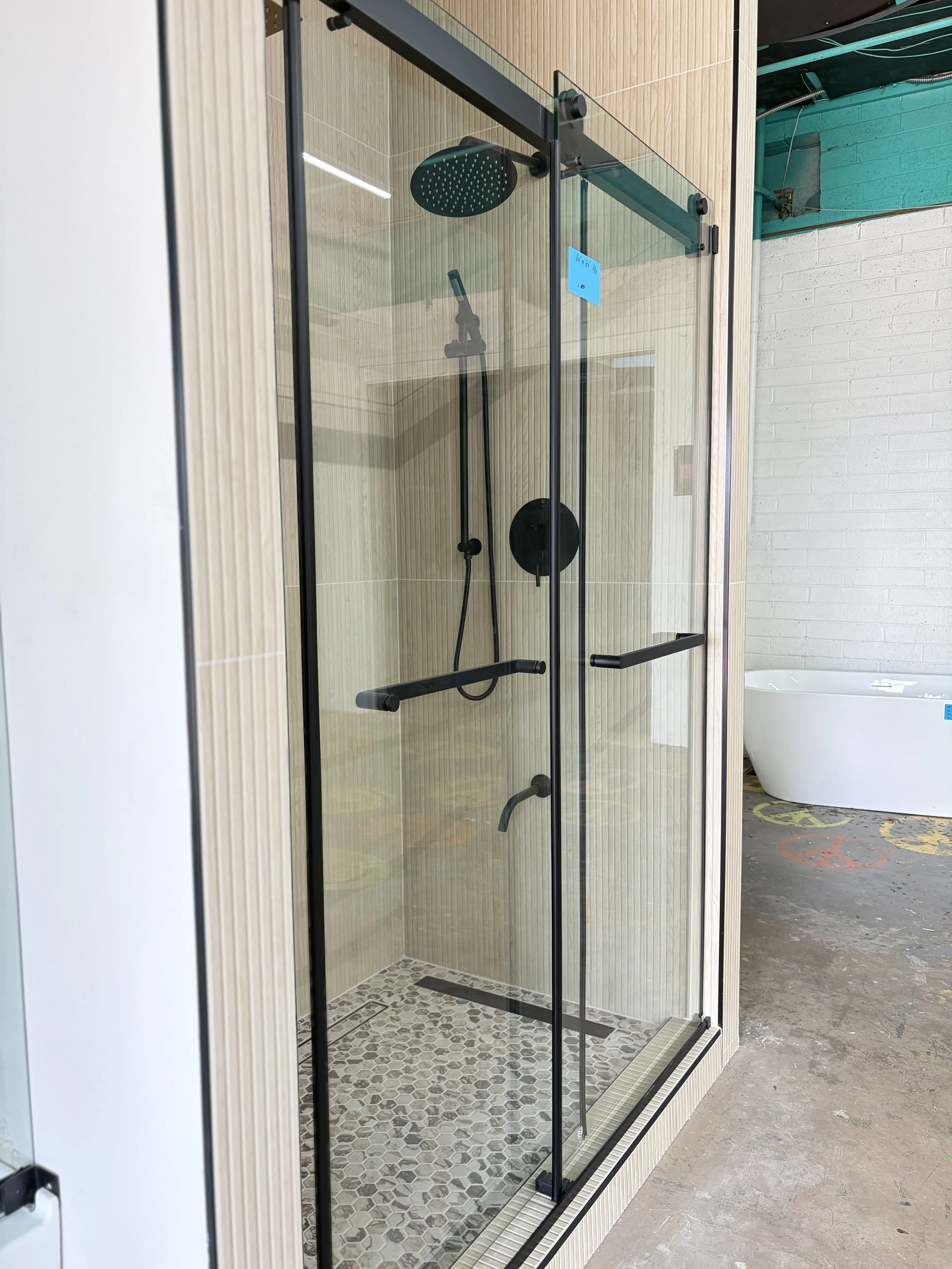 A modern shower enclosure with a glass door, black metal framing, a rain shower head, and a handheld shower head inside. The shower has beige tiled walls and a patterned tile floor. Surrounding areas include a white bathtub and a white brick wall, wi