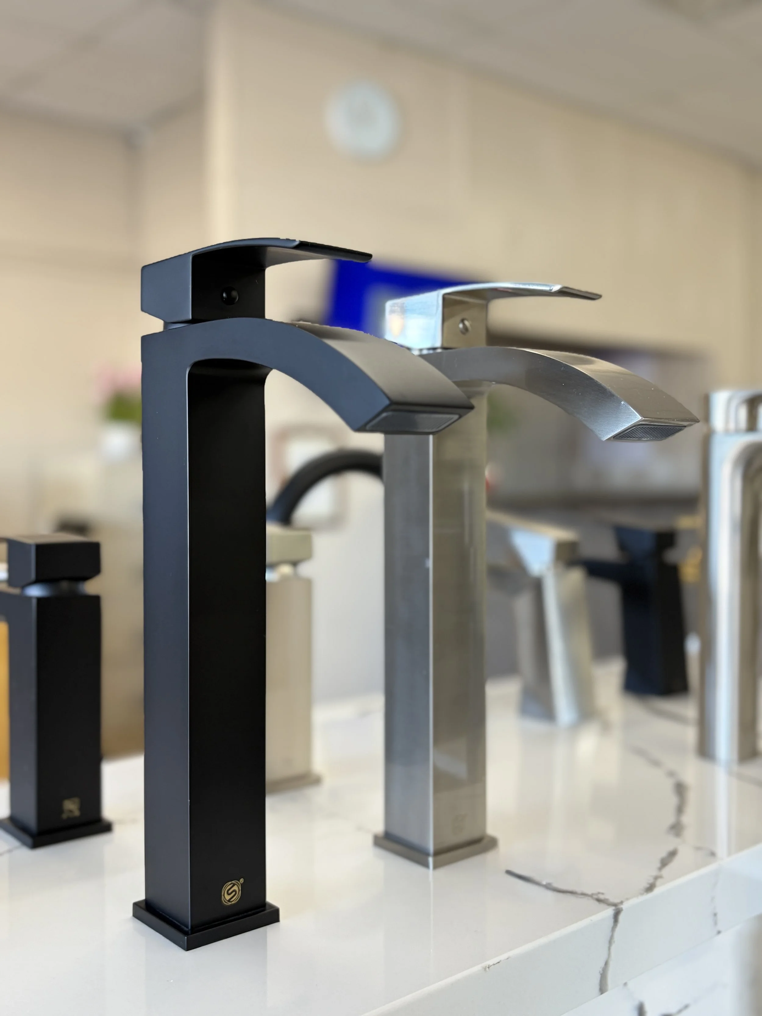A row of modern kitchen faucets in black, silver, and chrome finishes displayed on a white marble-like surface.