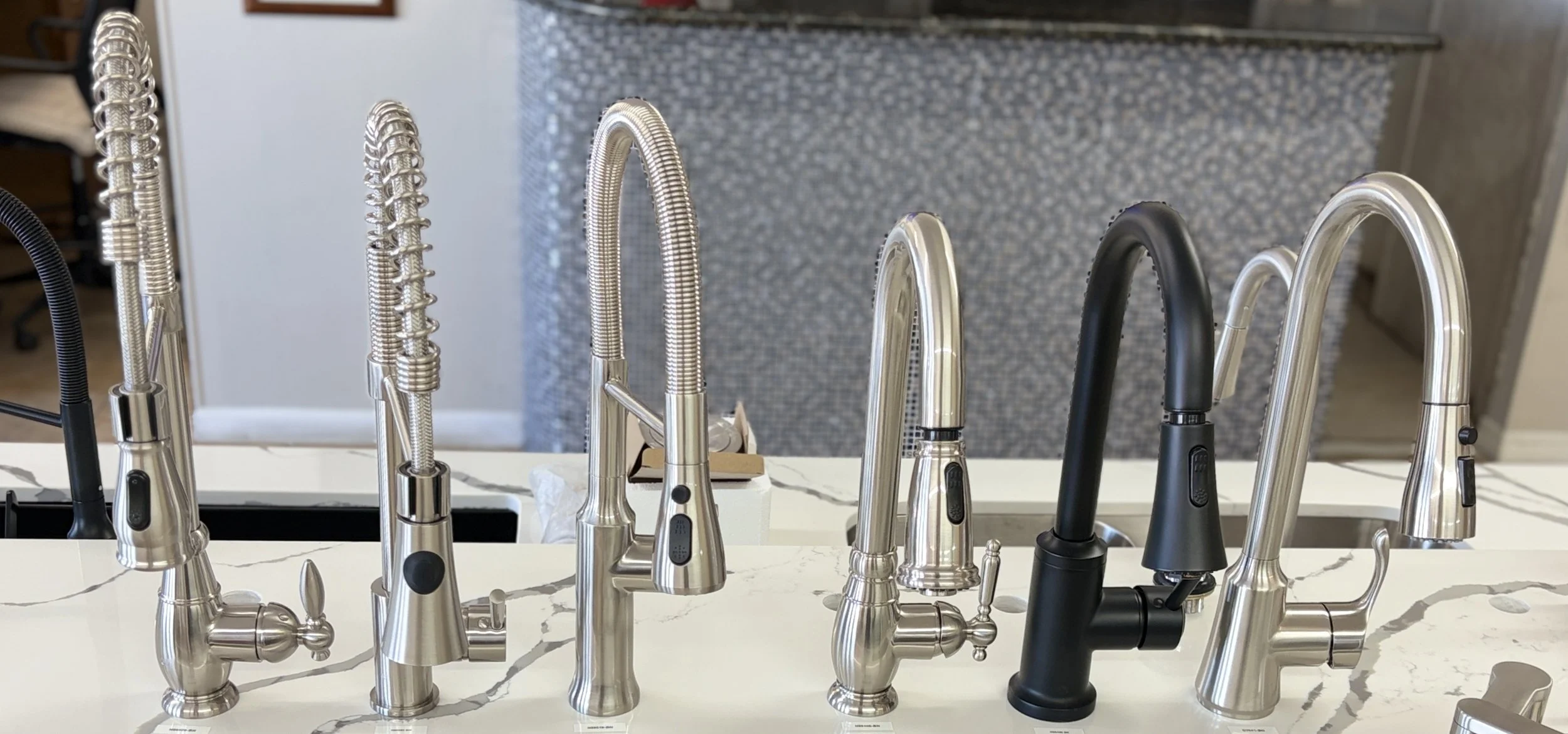 A display of various kitchen faucets in different styles and finishes on a marble countertop.