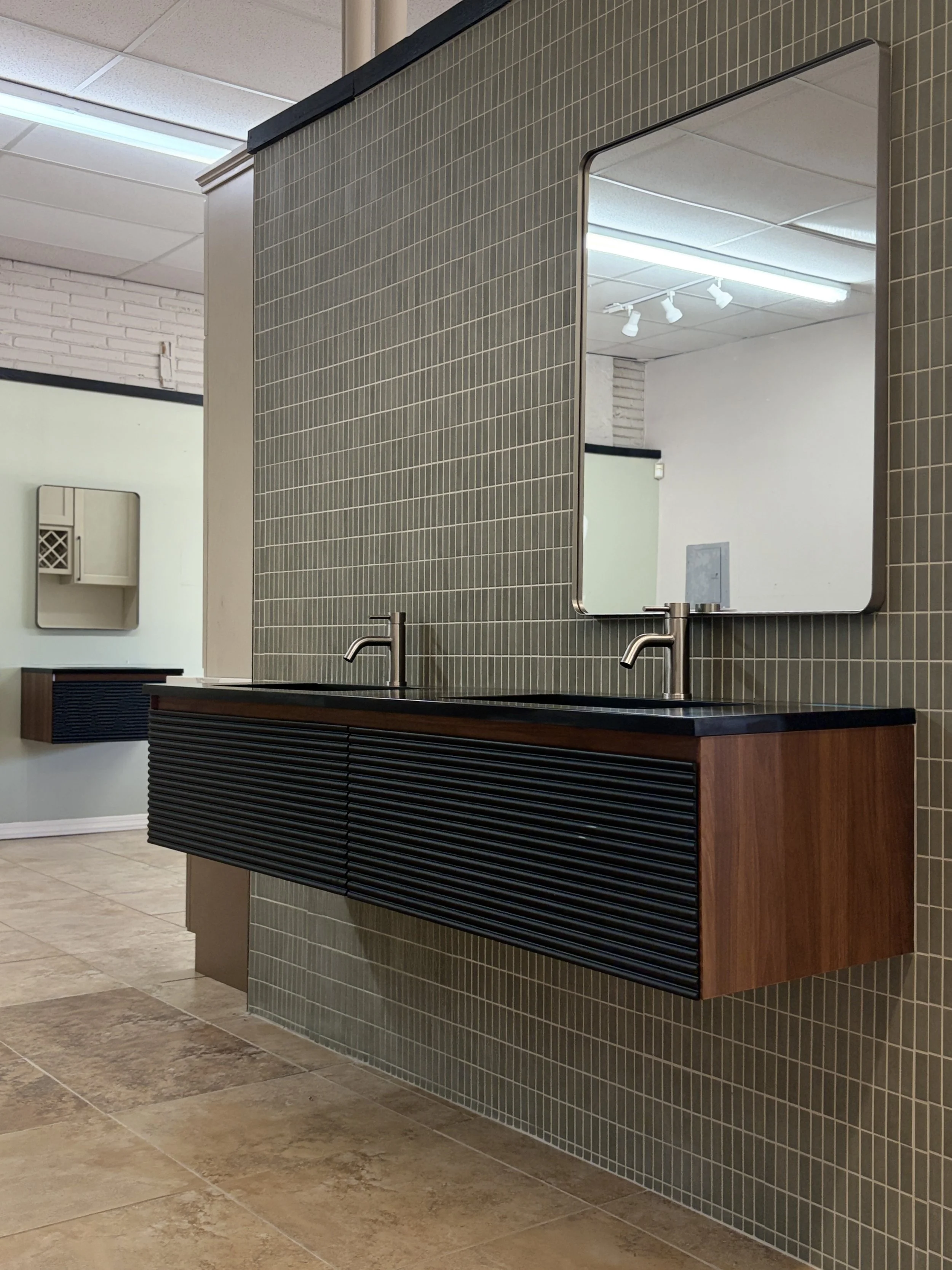 Bathroom sink area with a large mirror, dark wood cabinet, and modern plumbing fixtures. The walls are covered with small gray tiles, and the floor has beige tiles. Fluorescent and track lighting illuminate the space.