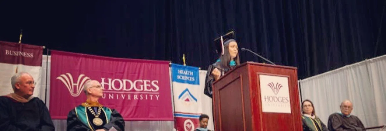 A woman in graduation gown and cap speaking at a podium during a commencement ceremony at Hodges University, with faculty members seated on stage behind her.