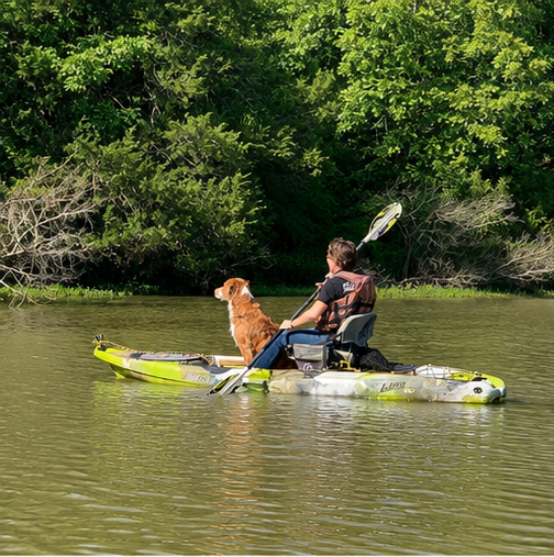 A woman and a brown and white dog sitting on a kayak in a river surrounded by lush green trees.