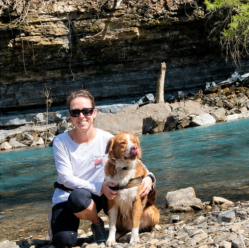 A woman wearing sunglasses and a white shirt kneels next to a brown and white dog beside a rocky riverbank with a cliff and trees in the background.
