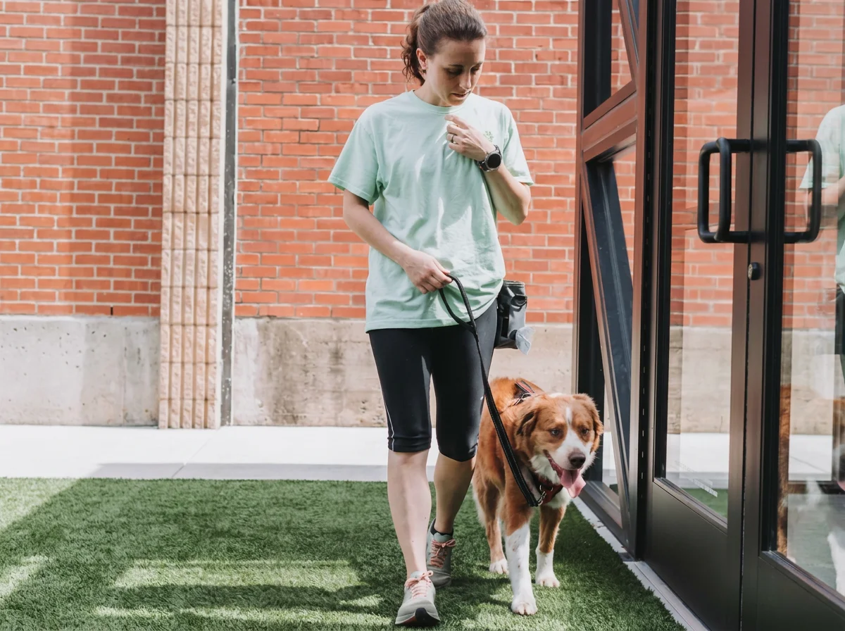 A woman in athletic clothing walking her dog on a leash outside a brick building entrance.