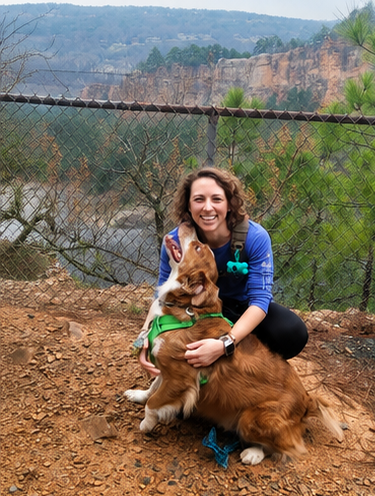 A woman crouching on dirt ground smiling while holding a large brown and white dog, outdoors with a fence, trees, and rocky cliffs in the background.