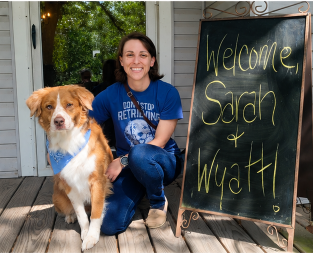 A woman with short brown hair, wearing a blue T-shirt and jeans, kneeling beside a medium-sized dog with a golden and white coat, on a wooden deck outside a home. There is a blackboard sign next to them that reads 'Welcome Sarah & Wyatt' in yellow chalk, welcoming Sarah and her dog Wyatt.