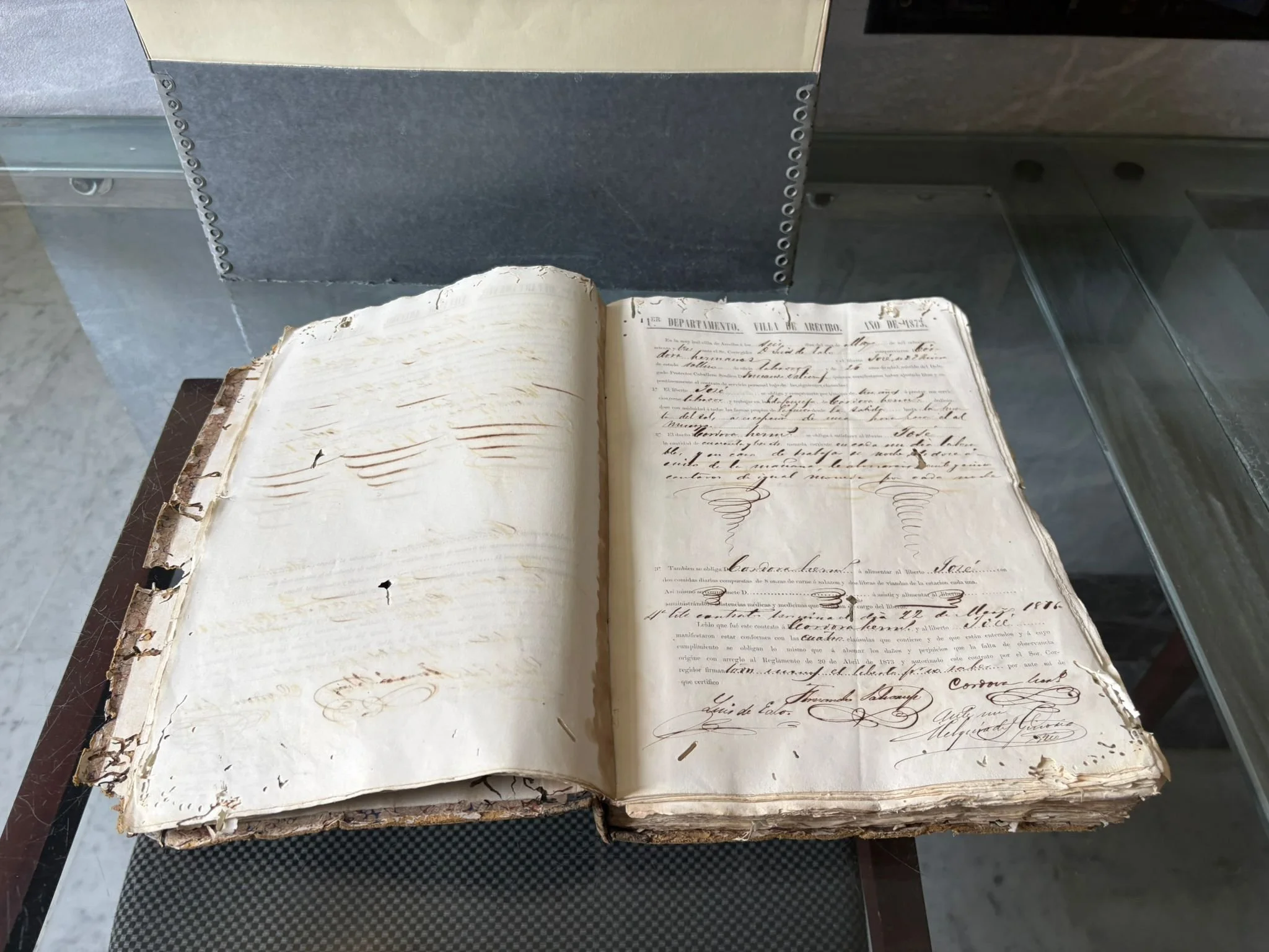 An old, weathered, handwritten manuscript or book with torn and damaged pages, resting on a surface with a transparent display case in the background.
