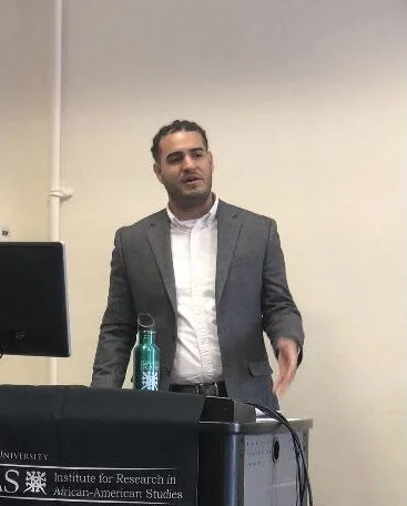 A man in a gray blazer and white shirt standing at a podium with a computer monitor, a water bottle, and a nameplate that reads 'Institute for Research in African-American Studies'.