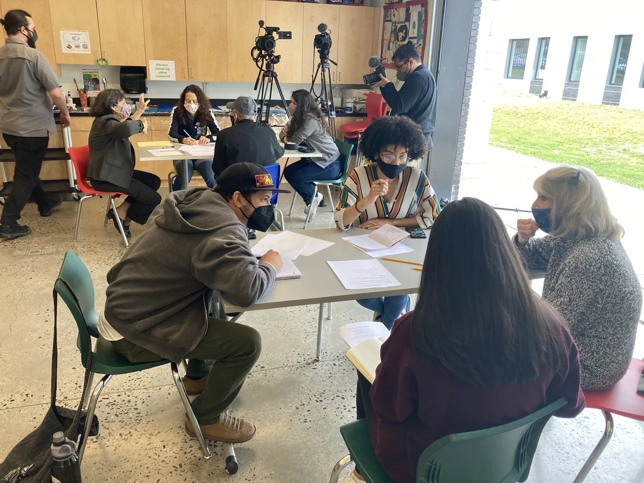 People sitting around a table in a room, engaged in a discussion, with cameras and videographers capturing the scene, and others working or observing nearby.
