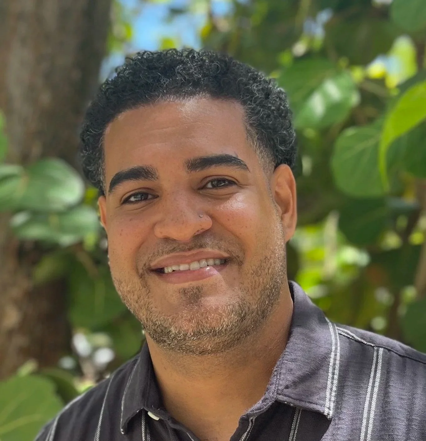 Close-up of a smiling man with curly black hair, wearing a dark striped shirt, standing outdoors with green leaves in the background.