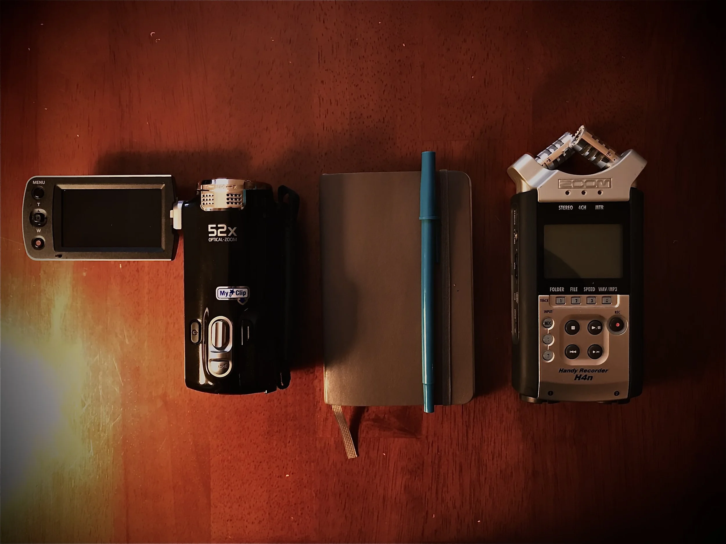 A wooden desk with a digital camera, a small black multimedia player, a closed notebook, a blue pen, and two digital recorders on it.