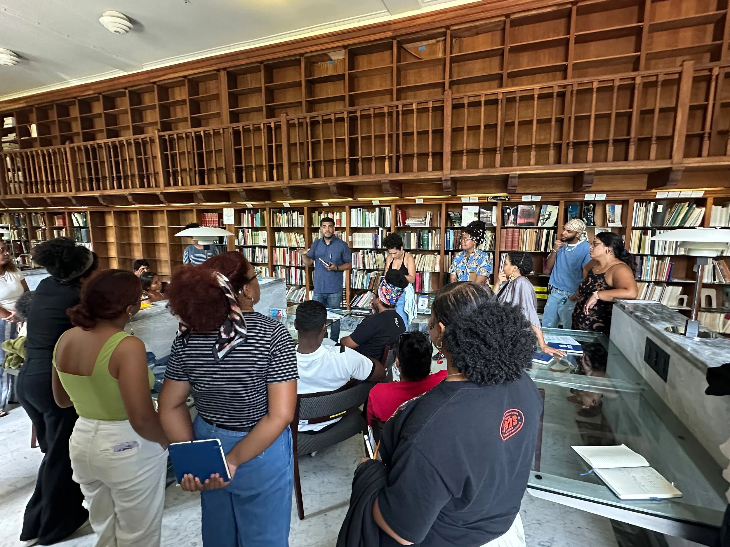 A group of people gathered in a library listening to a man holding a phone or tablet, with bookshelves in the background.