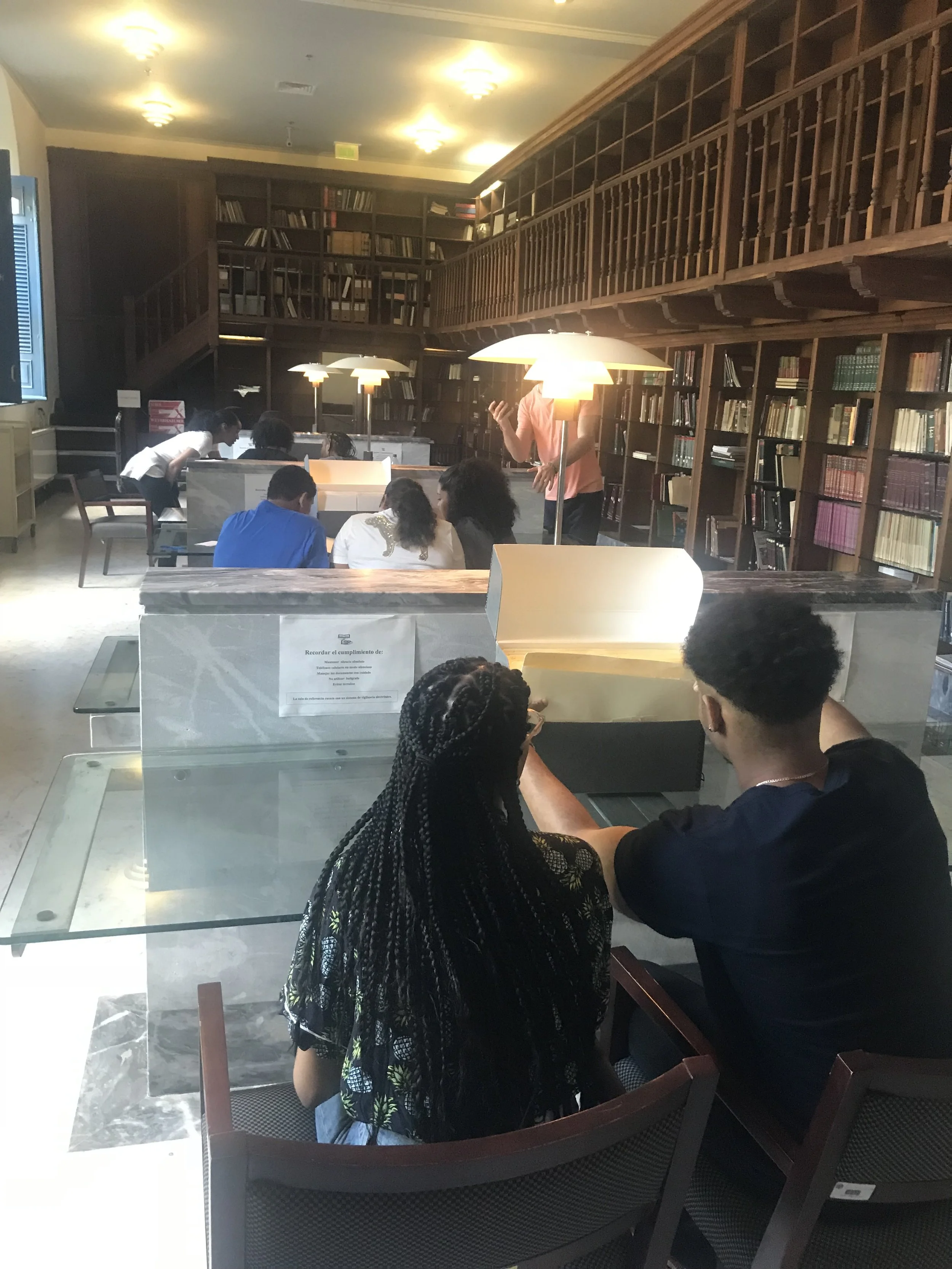 A library or study room with wooden bookshelves filled with books, some people sitting and chatting, and others standing around, illuminated by warm ceiling and table lamps.