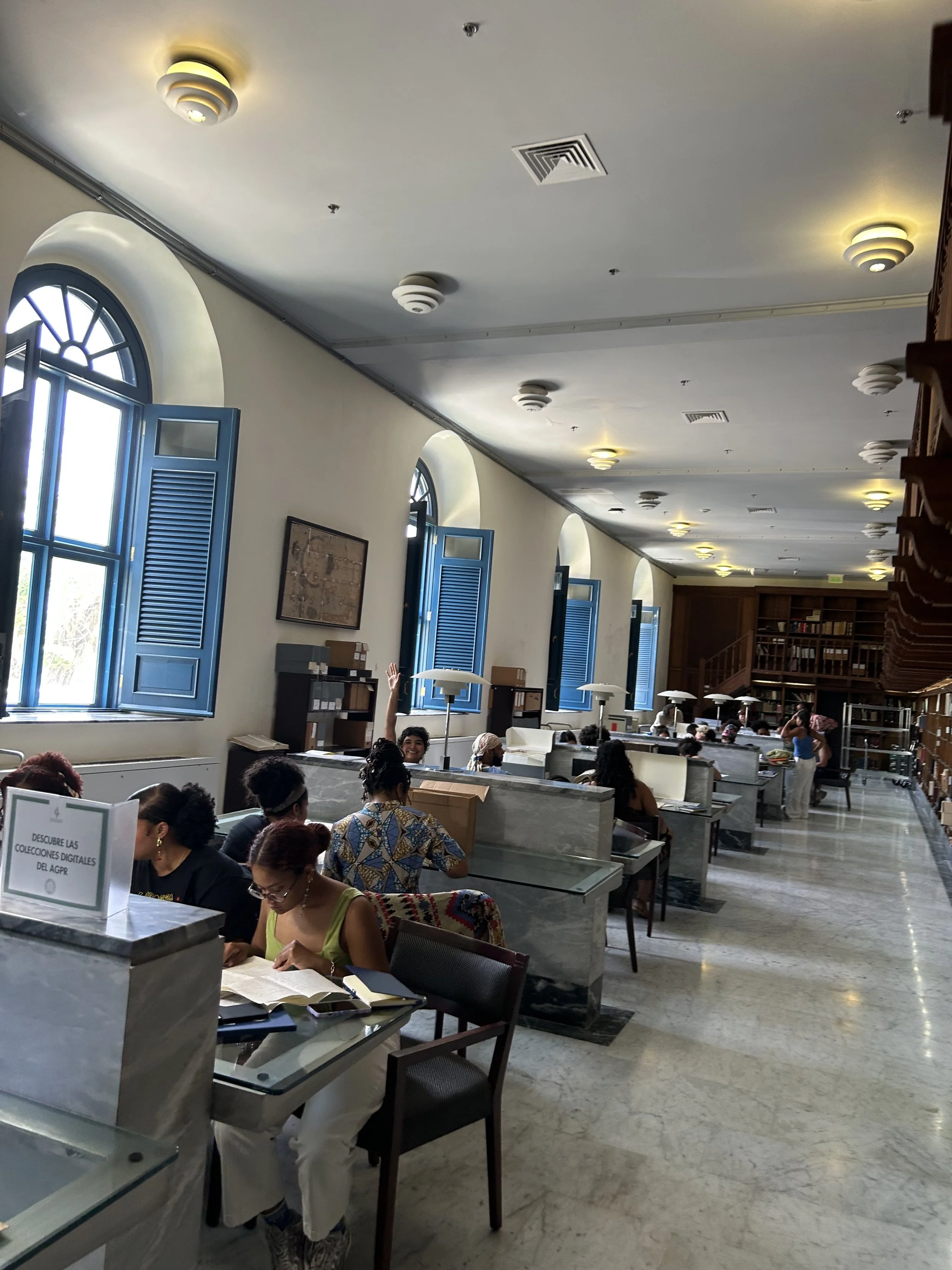 A large room with high ceilings, arched windows with blue shutters, and multiple ceiling lights. People sit at desks, some reading or writing, in a library or study space.