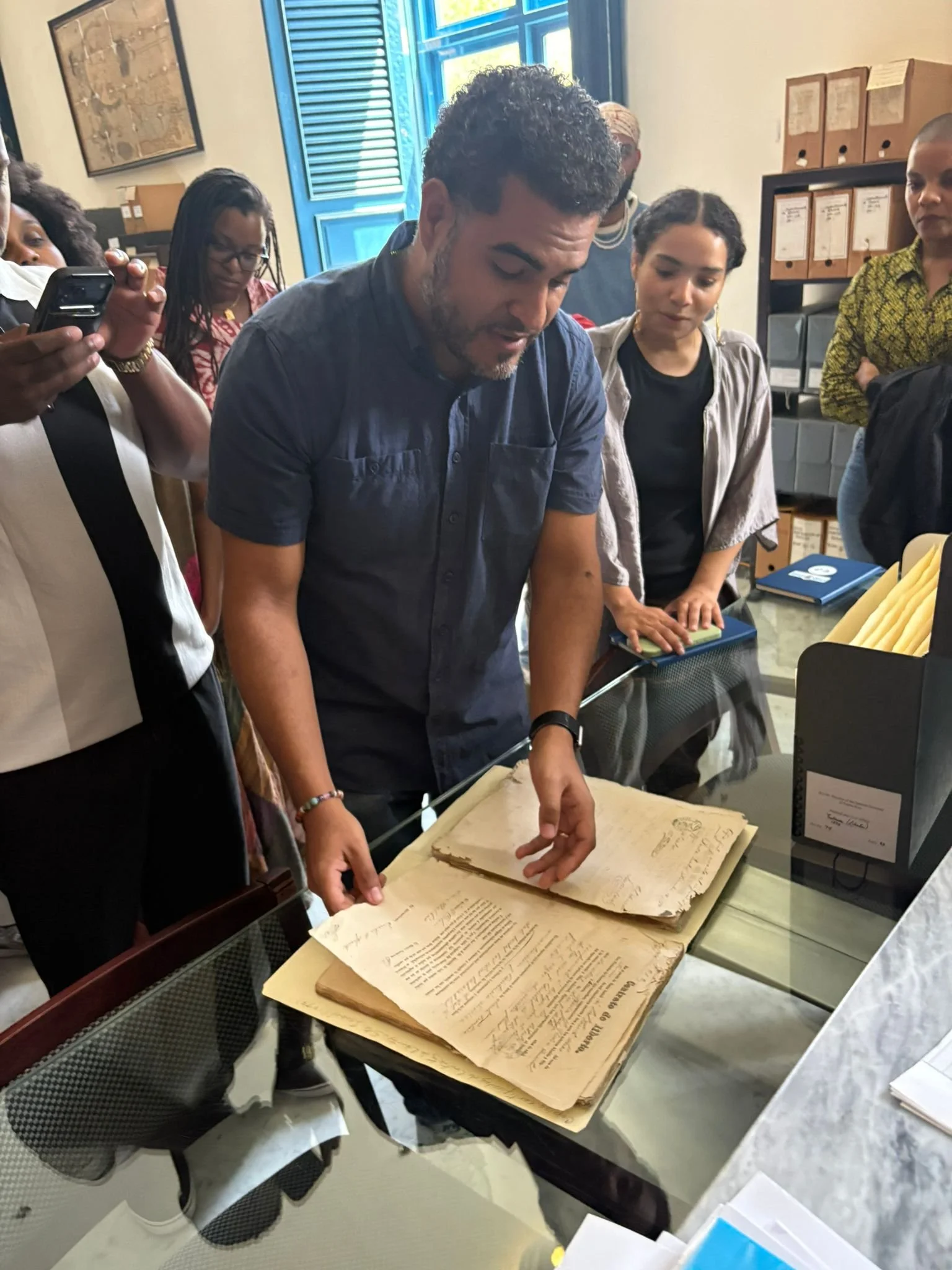 A group of people gathered around a table looking at ancient, large, handwritten documents or manuscripts. They appear to be in an office or archive room with shelves and boxes in the background. One person is pointing to the documents.