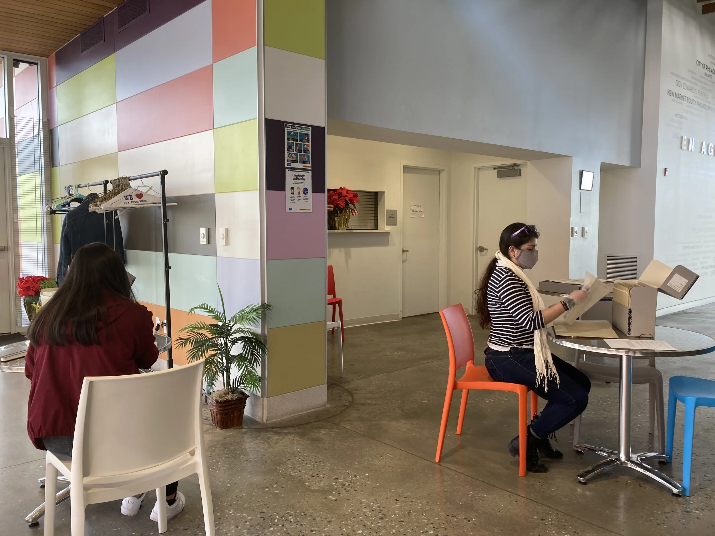 Indoor public space with colorful walls, people wearing masks, and a woman reading a paper at a table.