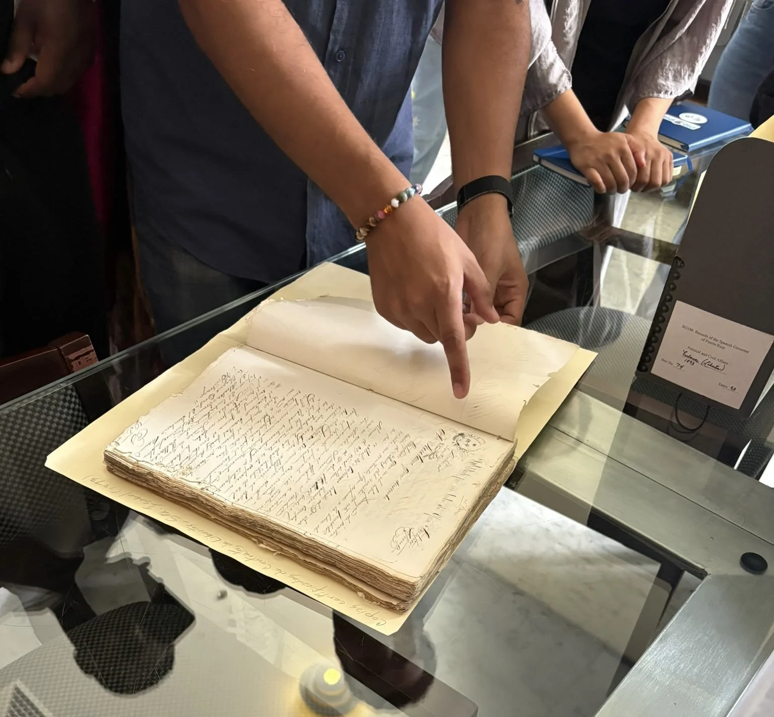 Hands pointing at an open old handwritten manuscript on display at a museum or archive.