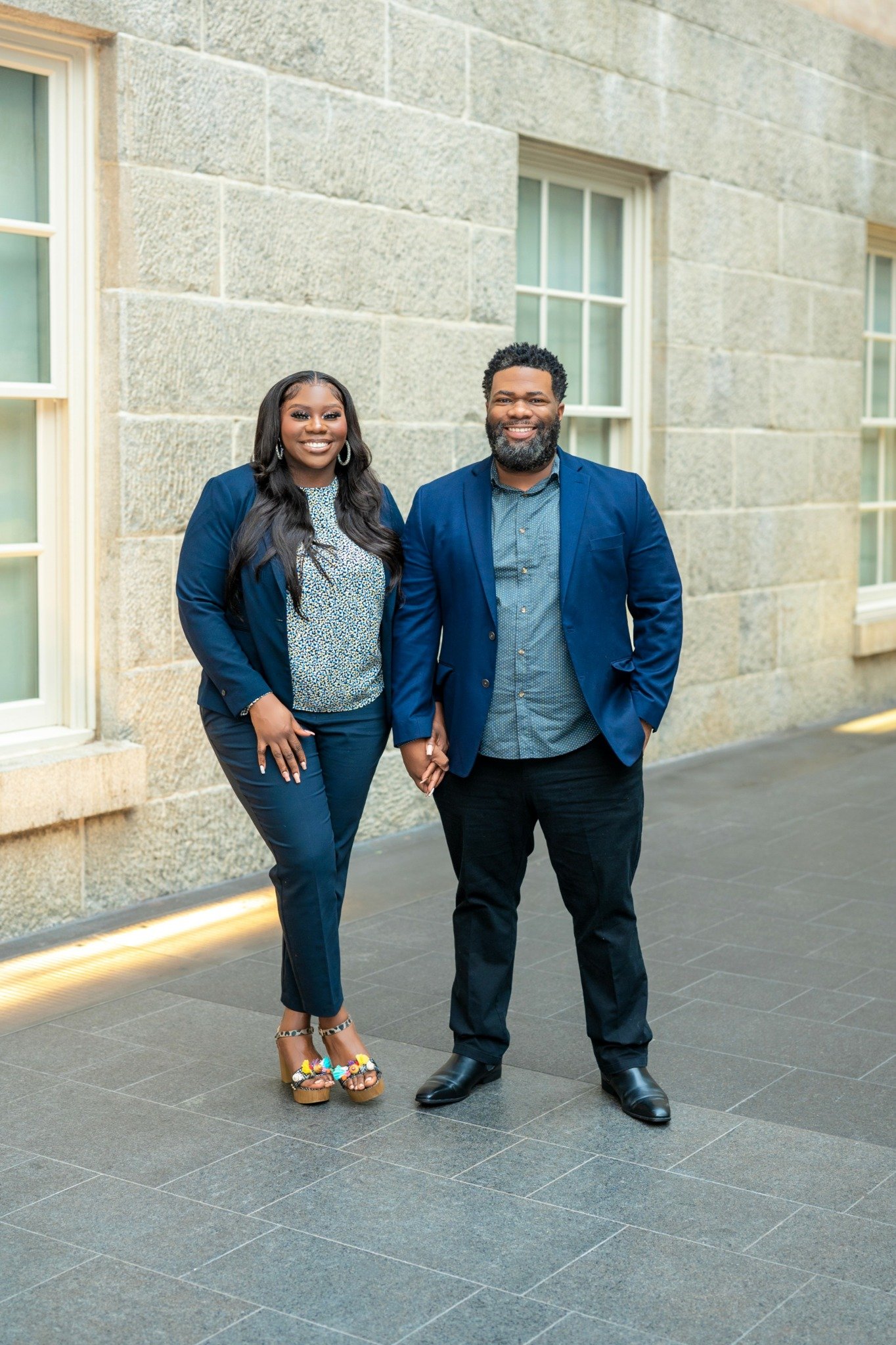 A smiling woman and man holding hands, standing outside near a stone building with large windows. The woman is wearing a blue blazer, patterned blouse, dark trousers, and colorful platform sandals. The man is dressed in a blue blazer, patterned shirt, and dark trousers with black shoes.