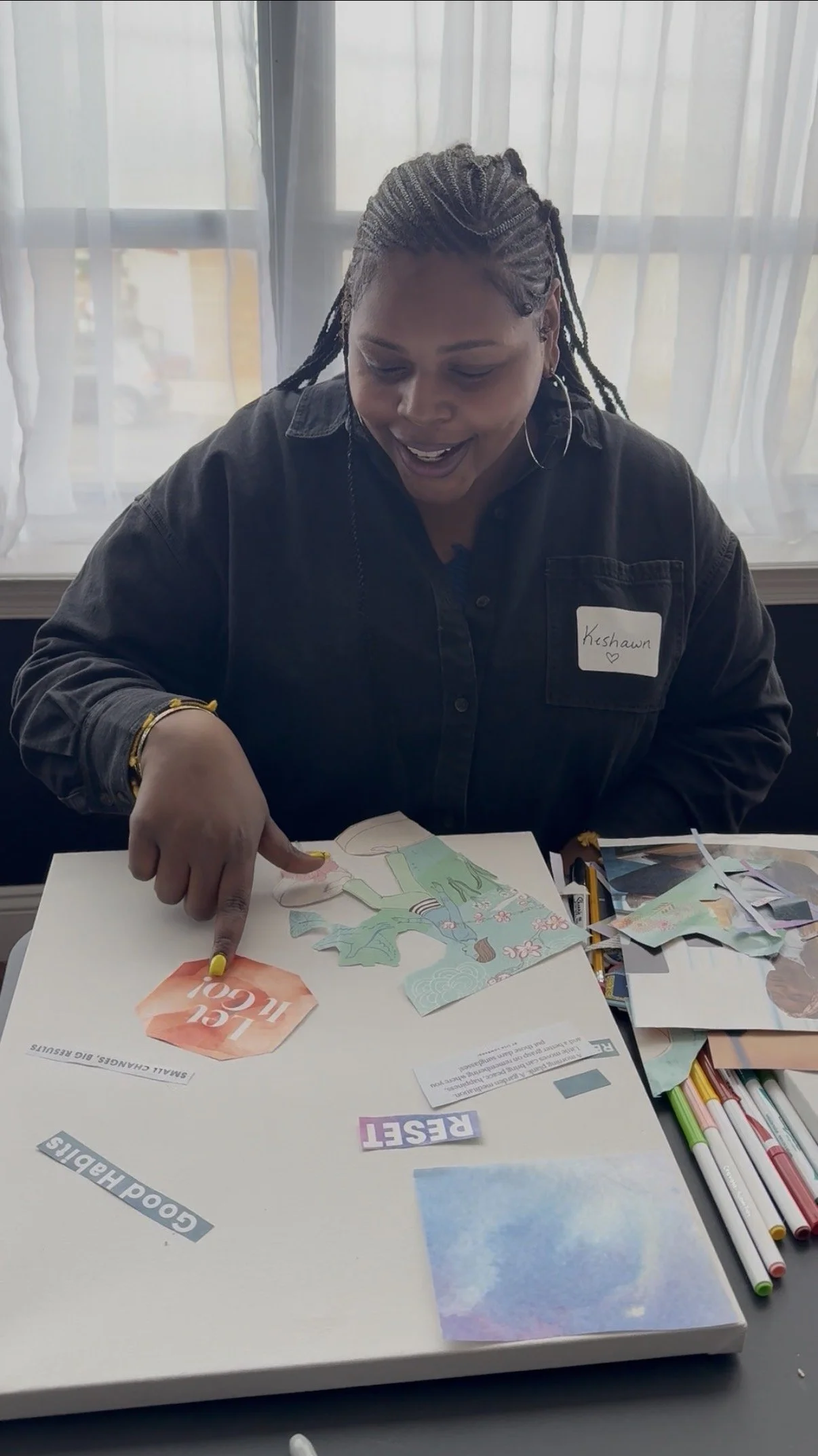 A woman with braided hair wearing a black shirt and hoop earrings is smiling and pointing at a puzzle with various cut out pieces and words on the table in front of her. The background has white curtains.