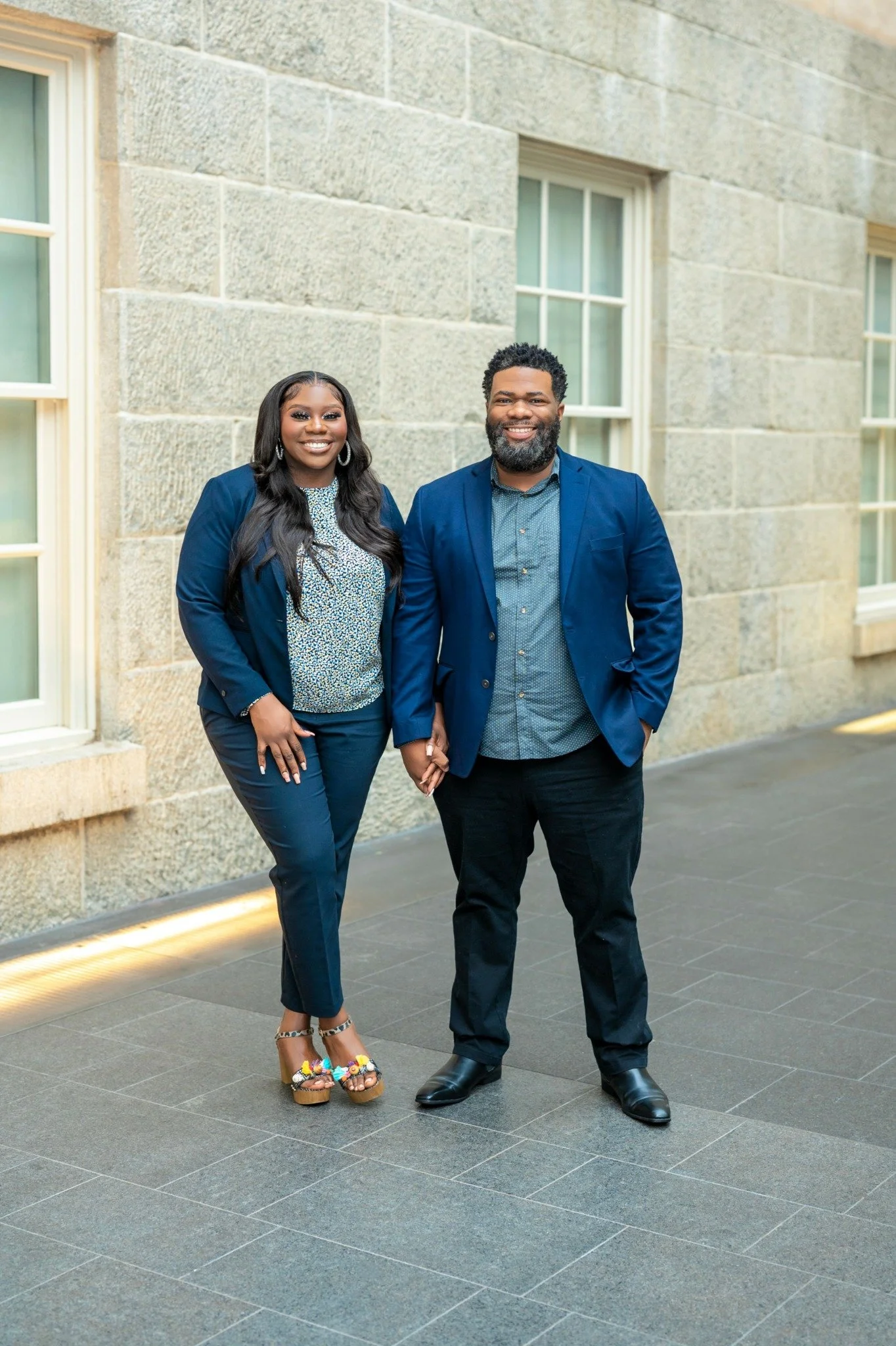 A smiling woman in a blue blazer and pants standing next to a smiling man in a blue blazer and dark pants. They are holding hands and standing on a sidewalk in front of a stone building with large windows.