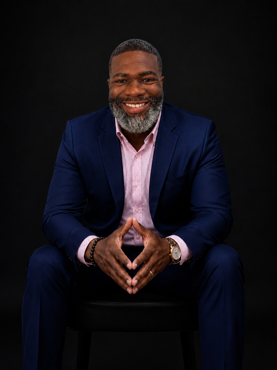 A smiling middle-aged man with a gray beard, wearing a navy blue suit and light pink dress shirt, sitting on a black stool against a black background, with his hands joined in a triangle shape.