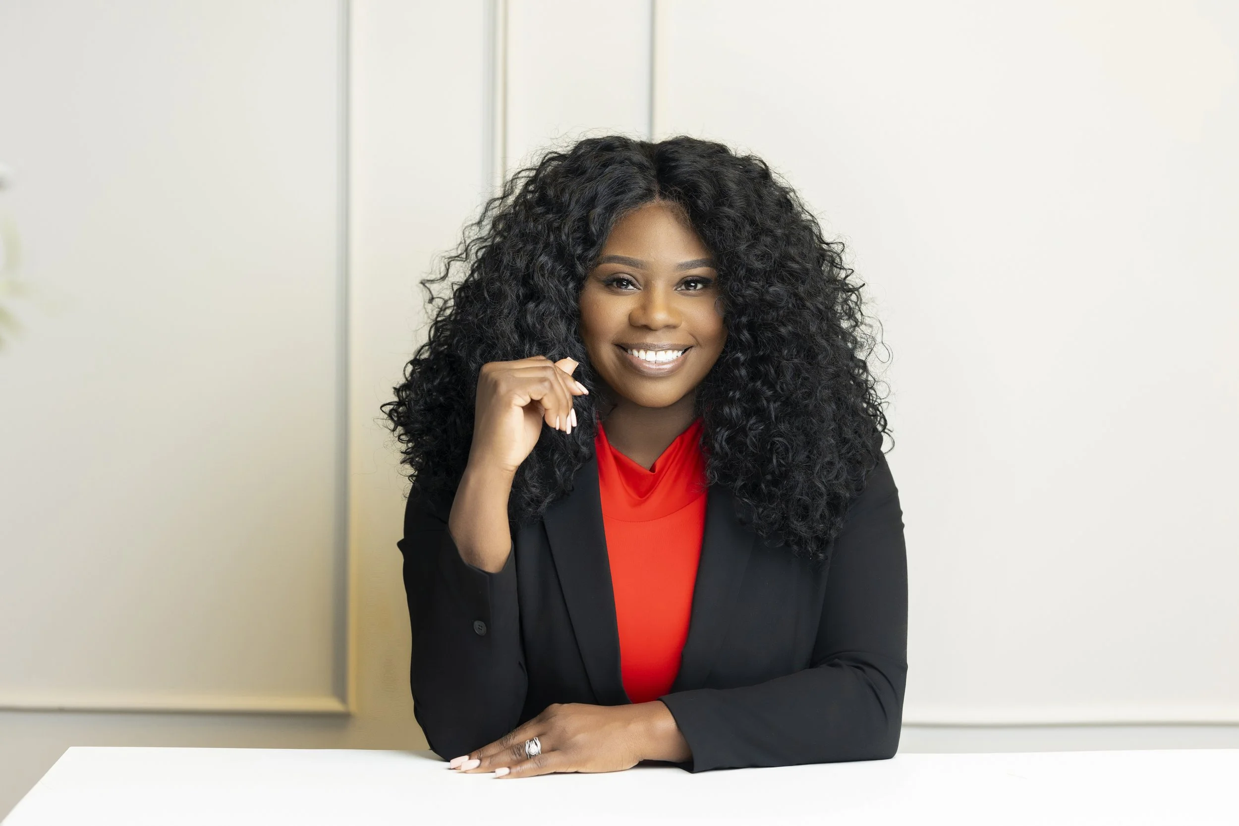 A woman with long curly black hair, wearing a red top and a black blazer, is sitting at a white table in front of a plain white wall. She is smiling and looking directly at the camera with her right hand resting on her chin.