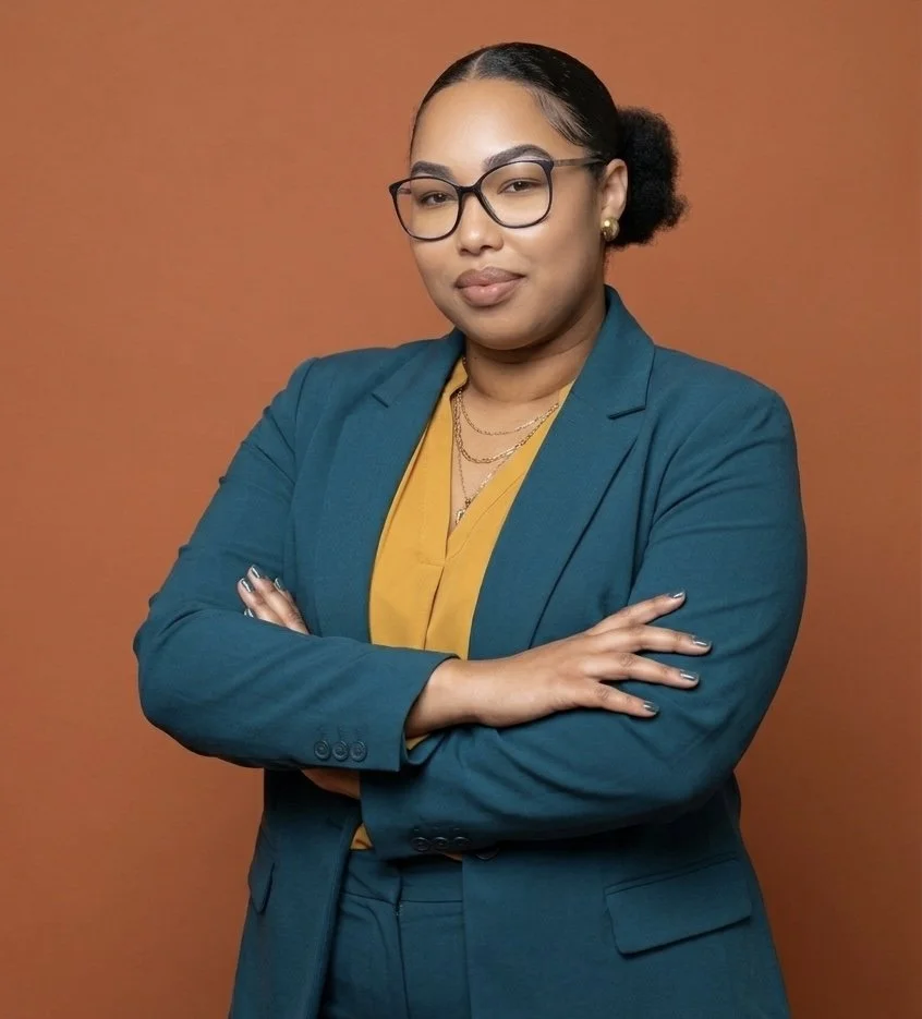 A confident woman dressed in a teal blazer and yellow blouse, wearing glasses, jewelry, and with arms crossed against an orange background.