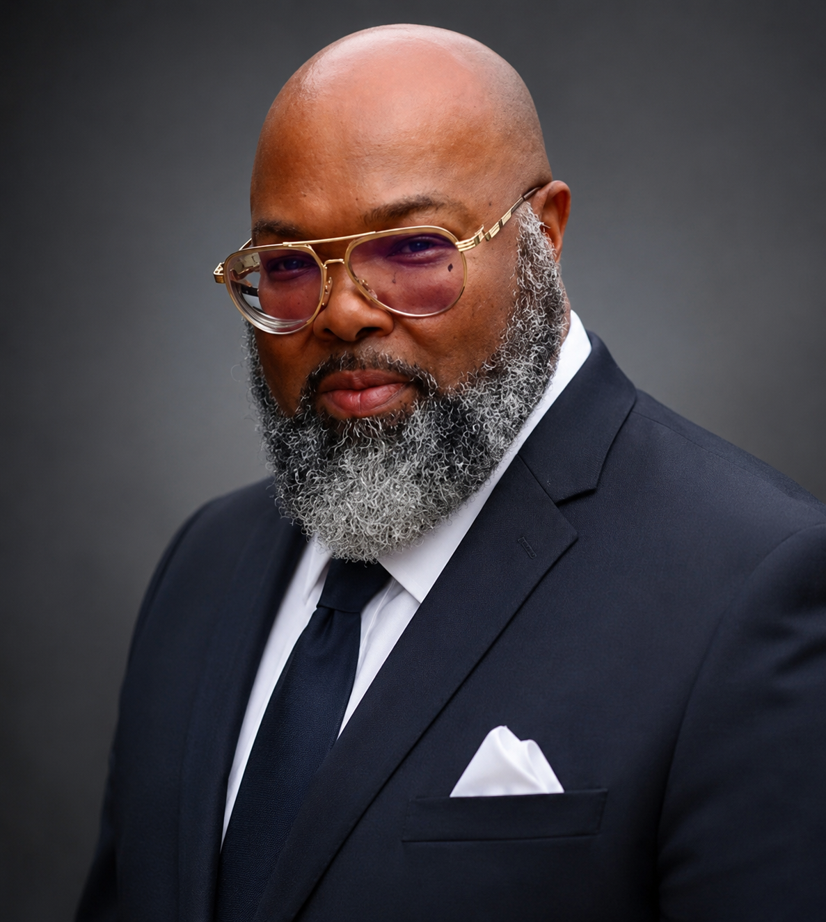 A bald man with a gray beard wearing glasses with pink-tinted lenses, dressed in a formal black suit, white shirt, and black tie, against a dark gray background.