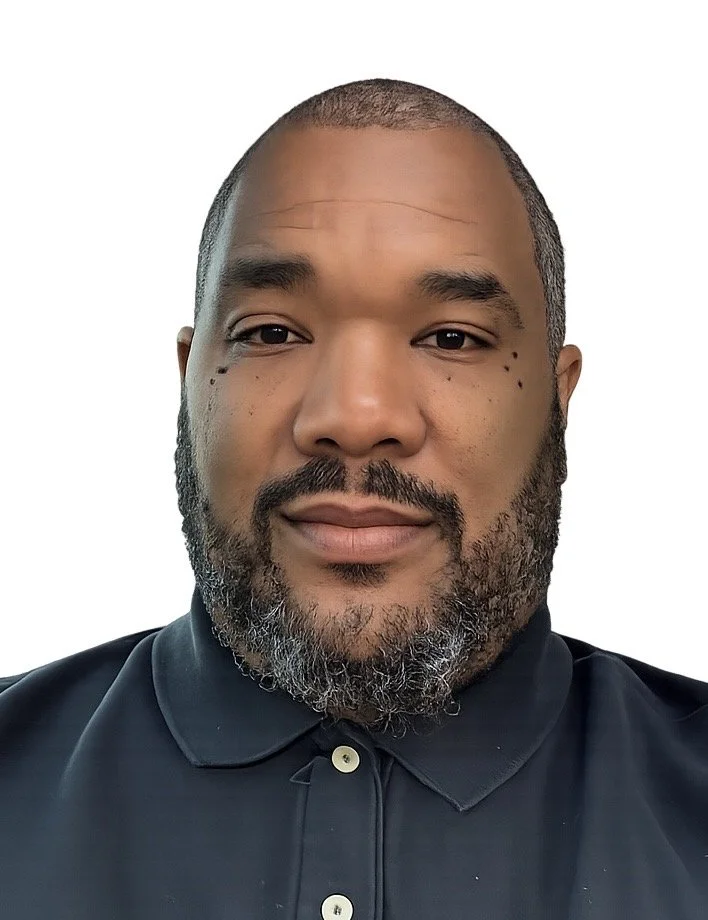 A close-up portrait of a man with a beard and short hair, wearing a dark collared shirt, against a plain white background.