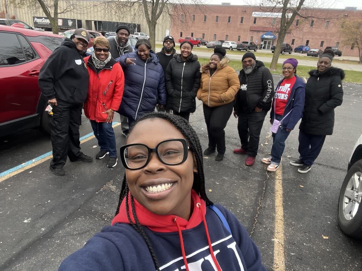 A group of ten diverse people posing for a selfie in a parking lot, smiling, with a young woman in the foreground wearing glasses and a navy hoodie taking the photo.