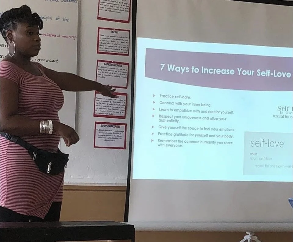 A woman giving a presentation in a classroom, pointing to a slide titled "7 Ways to Increase Your Self-Love". The slide lists tips such as practicing self-care, connecting with one's inner being, and practicing gratitude.