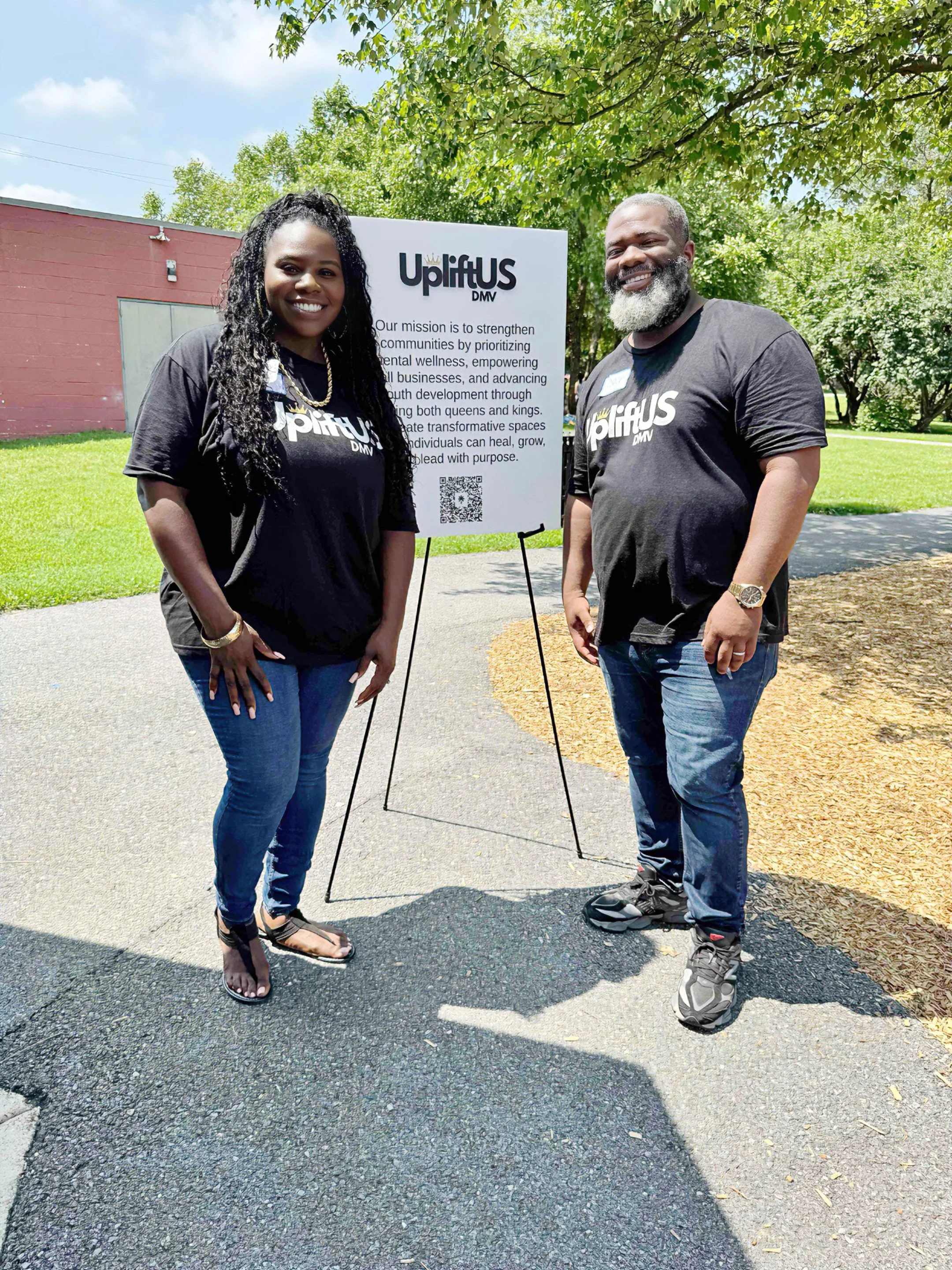 Two people standing outdoors, smiling, next to a sign for UpliftUS DMV, with trees and a red building in the background.