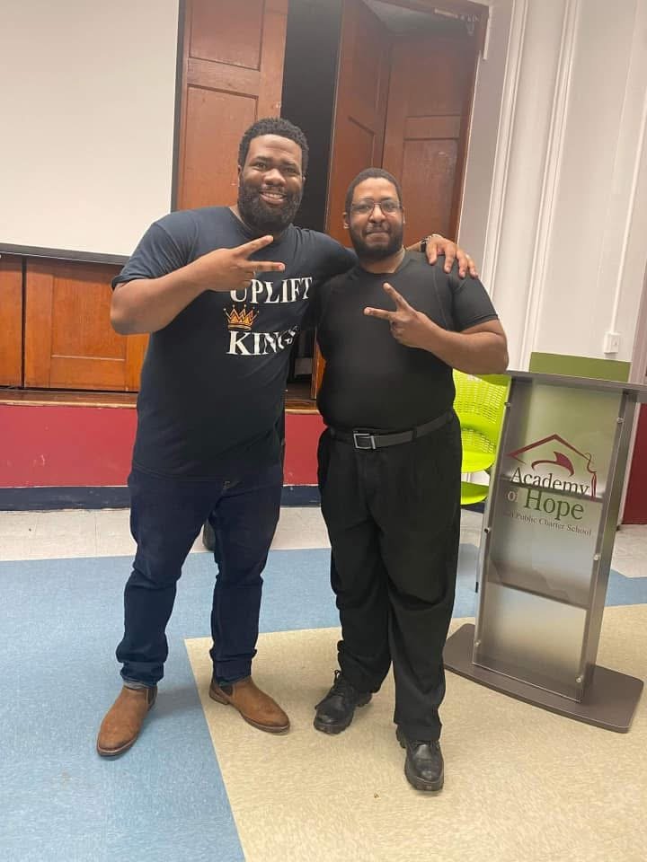 Two men standing together indoors, smiling, making peace signs with their hands. One wears a black t-shirt with 'Uplift Kings' logo, and the other wears a black shirt and pants. There is a sign that reads 'Academy of Hope' in the background.