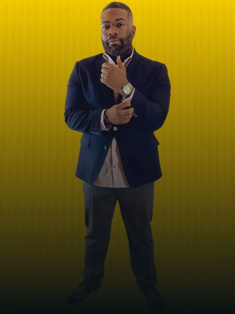 A man in a dark suit and patterned shirt standing against a yellow background, with one hand adjusting his collar and the other on his wristwatch.