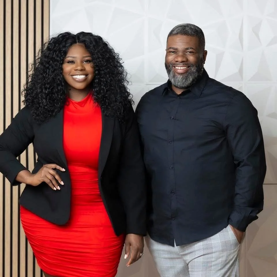 A woman with curly black hair wearing a black blazer and red dress standing next to a man with short gray hair and beard wearing a black shirt and gray plaid pants, both smiling at the camera.