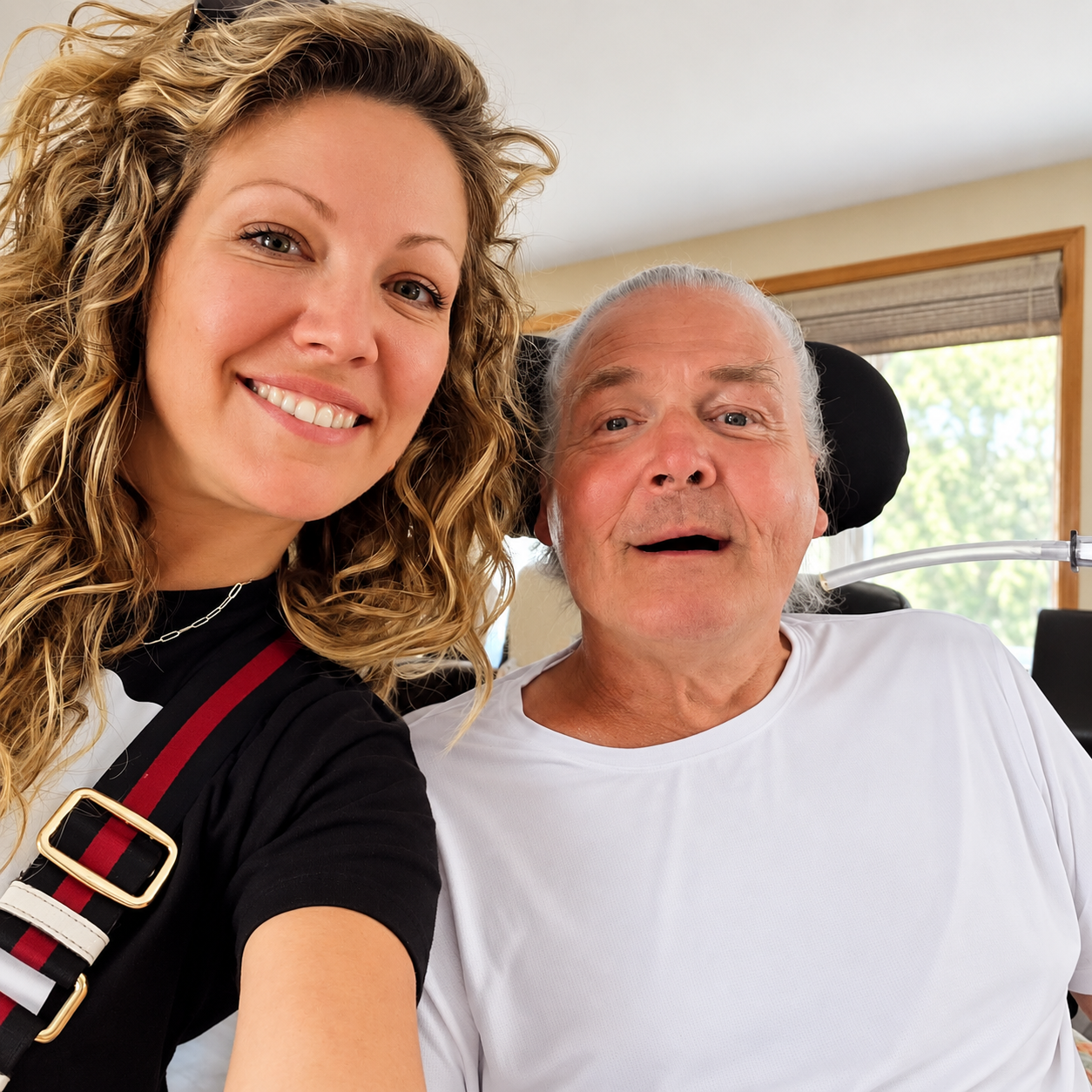 A woman with curly blonde hair smiling and taking a selfie with an elderly man in a wheelchair, inside a room with a window showing green trees outside.