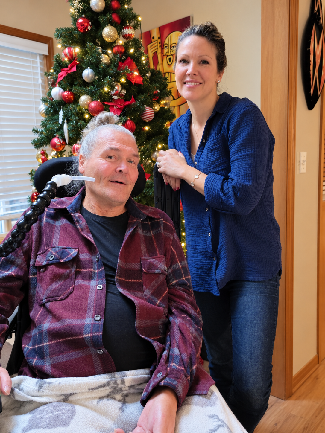 A man with a disability sitting in a chair with a Christmas tree in the background, accompanied by a woman. The man is wearing a plaid shirt over a black shirt and has a device near his neck. The woman is standing beside him, smiling, and wearing a blue shirt and jeans.