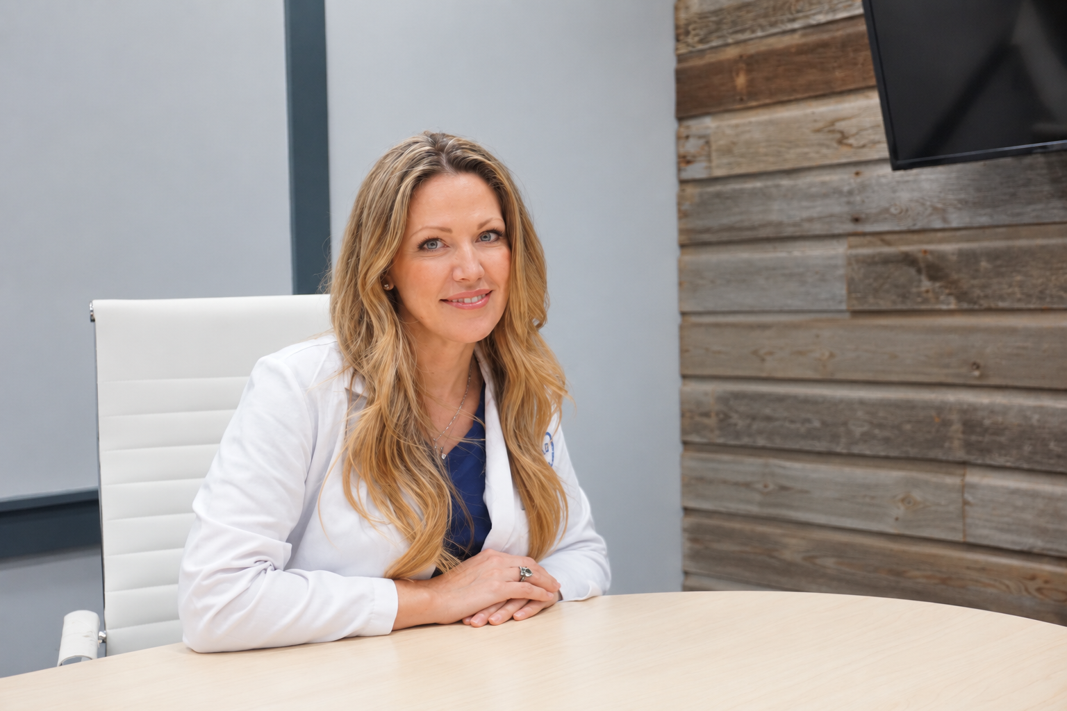 A woman sitting at a conference table in an office, smiling, with a wooden wall and flat screen monitor in the background.