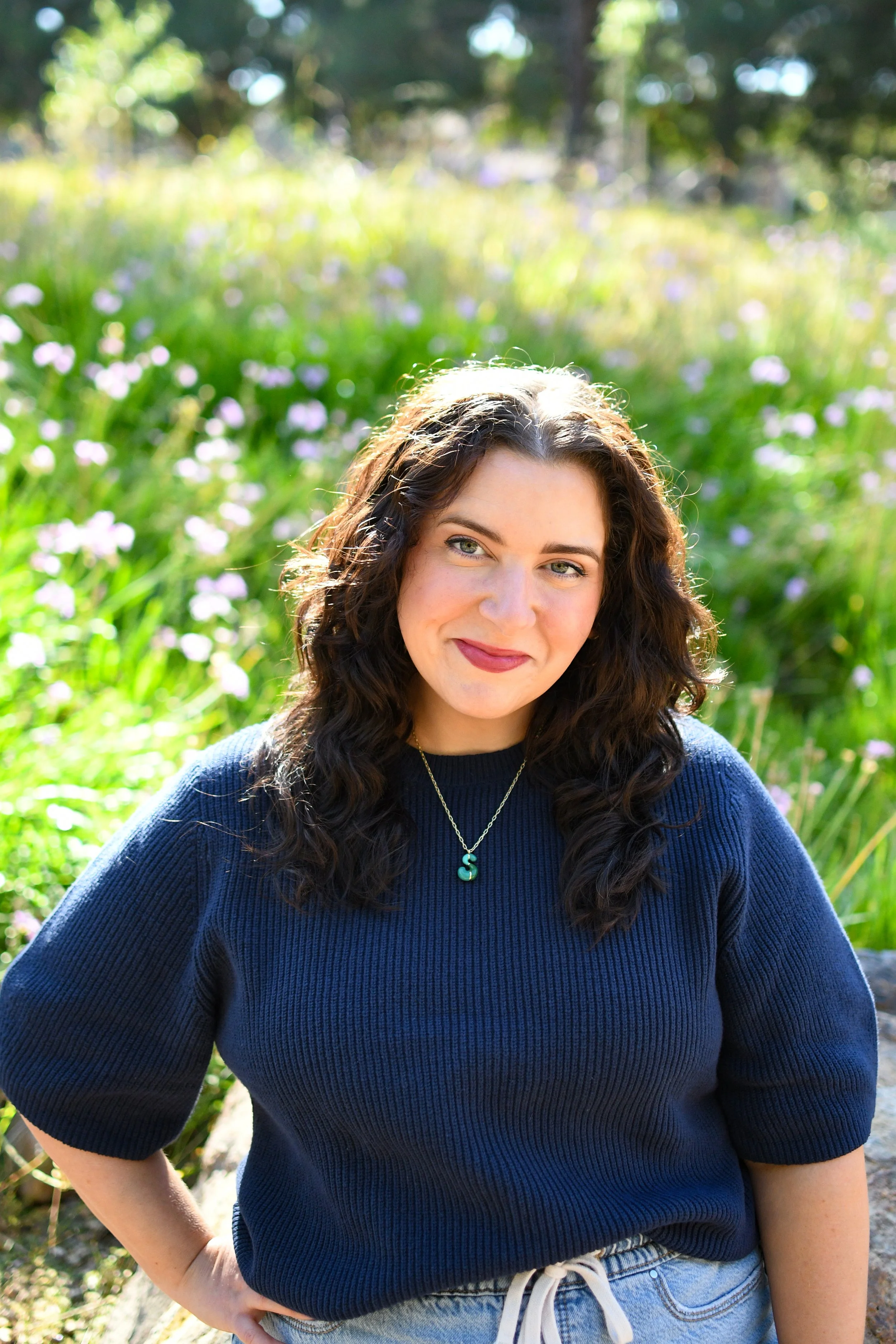 A woman with curly dark hair and light skin smiling outdoors, wearing a navy blue sweater and a necklace with an "S" pendant, standing in a sunlit garden with green foliage and purple flowers in the background.