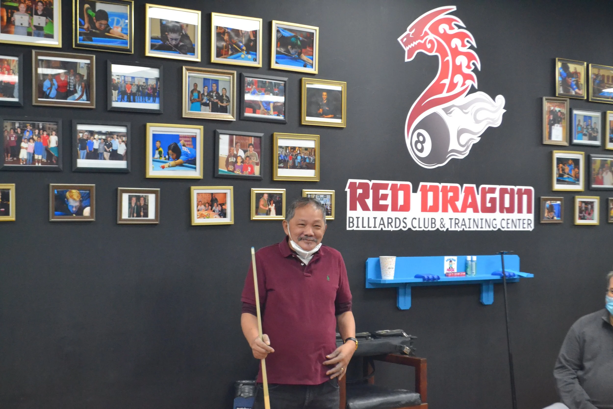 Indoor scene featuring a man in a maroon shirt holding a pool cue, standing in front of a wall decorated with multiple framed photographs of people playing billiards, tournament moments, and group images. The wall also displays a logo for the Red Dragon Billiards Club & Training Center.