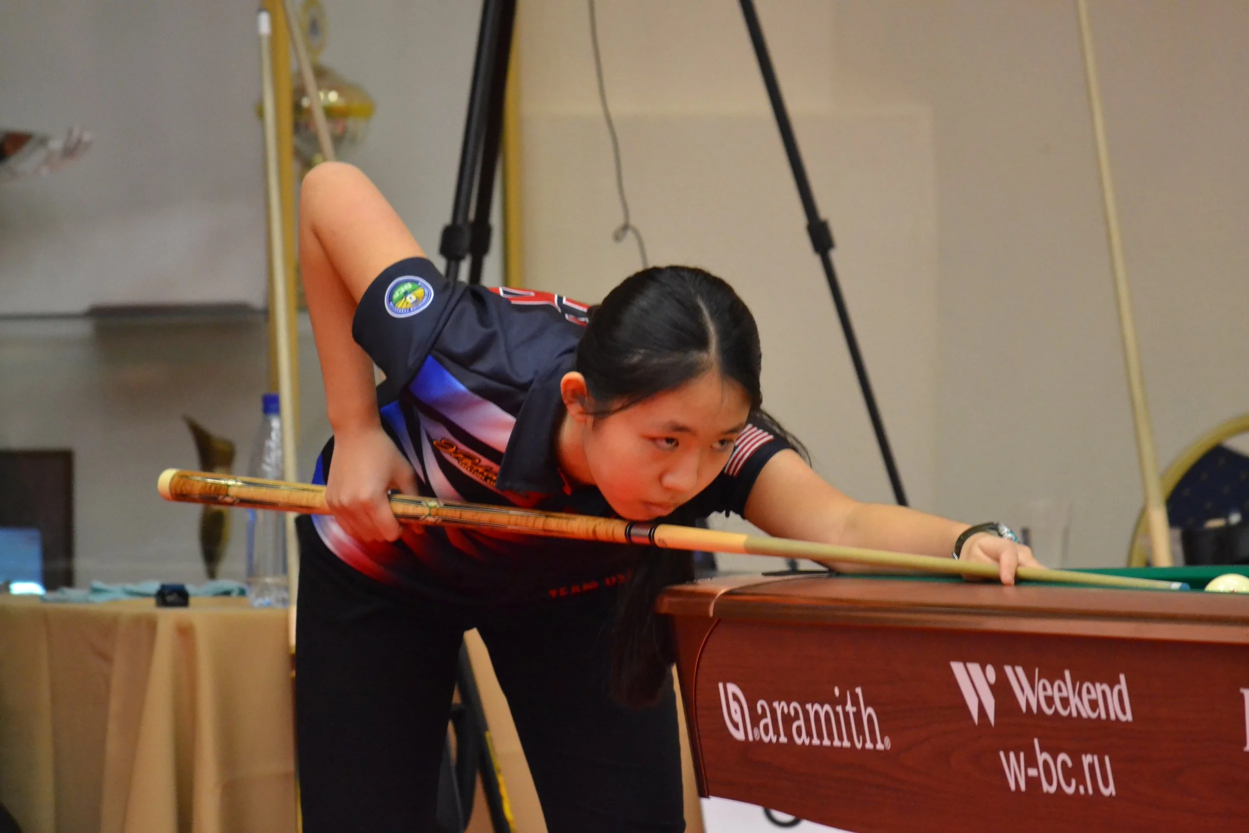 A woman playing pool, leaning over the table with a cue stick, aiming to make a shot. She has dark hair tied back and is wearing a black and red sports shirt. The scene appears to be indoors, possibly at a sporting event or recreational venue, with tables and banners in the background.